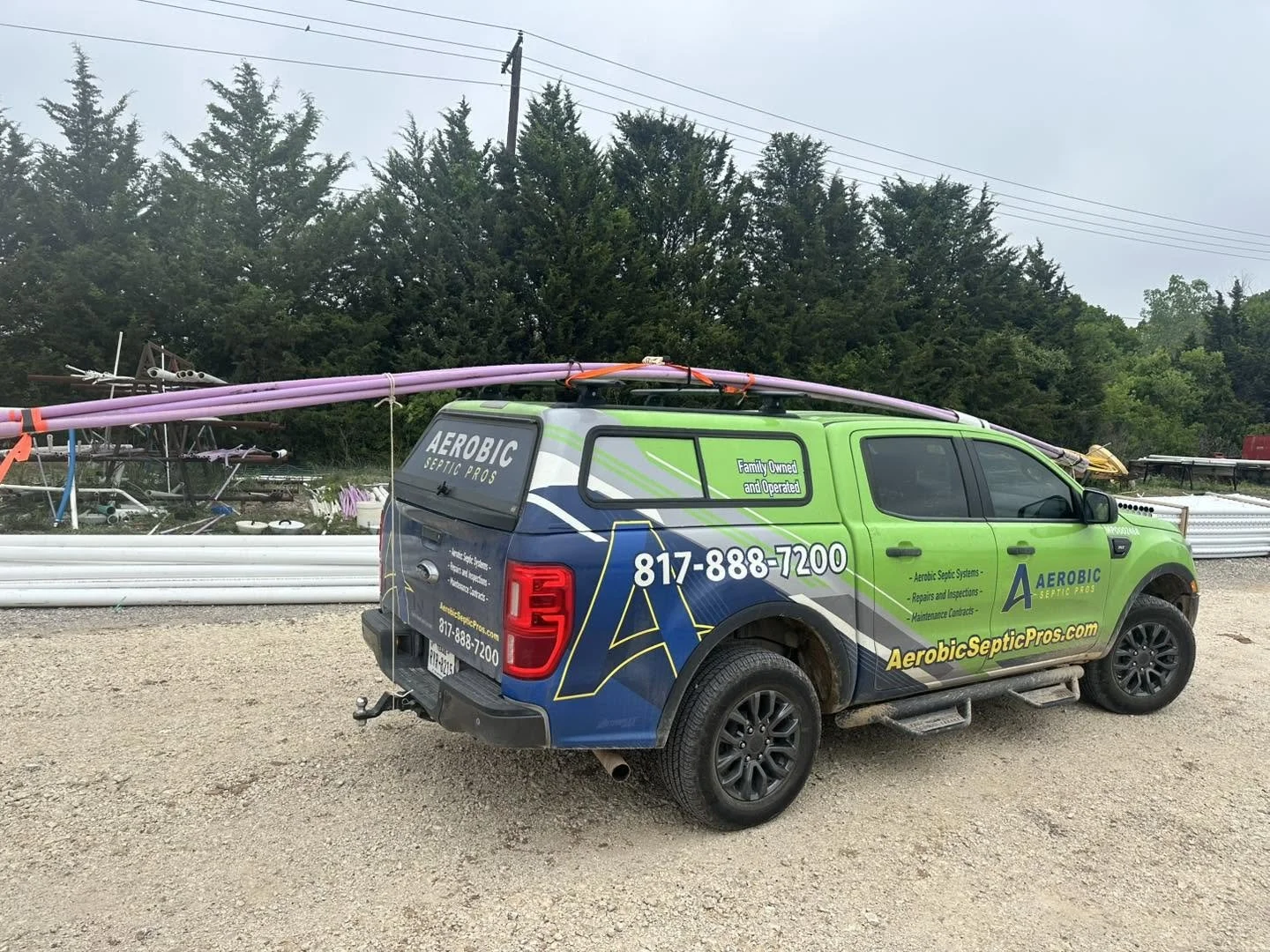 A green and blue company pickup truck with the logo 'Aerobic Septic Pros' is parked on a gravel lot, with purple pipes and septic equipment in the background.