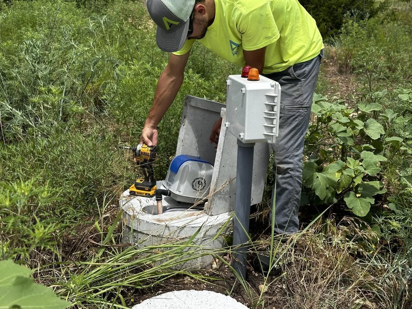 A man in a neon yellow shirt and gray pants is working on a sewer access point in a grassy outdoor area, using a cordless drill, with various tools nearby.