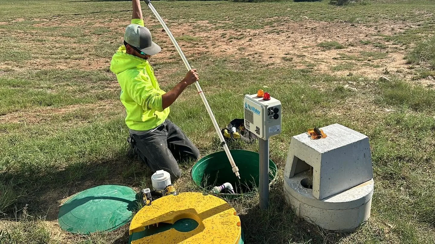 A person kneeling in an outdoor grassy area, wearing a bright yellow hoodie, a cap, and dark work pants, working with water monitoring equipment next to a water test well and a concrete water access point.
