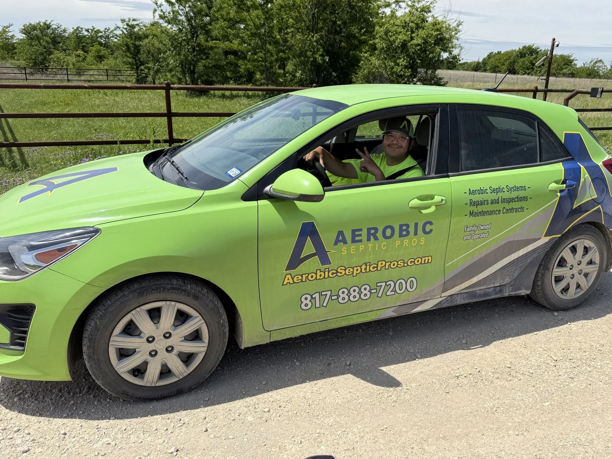 A green car with company branding for Aerobic Septic Pros parked on a dirt road, with a man smiling and making a peace sign inside. The background features trees, a metal fence, and a cloudy sky.