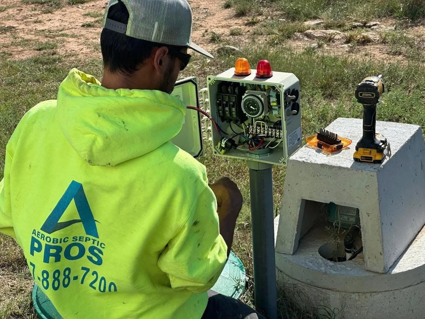 A person wearing a yellow high-visibility jacket and a green cap is working on an electrical control box outdoors. The control box is mounted on a stand, and the person is using tools to work inside it. There is a cordless drill and a small toolbox w
