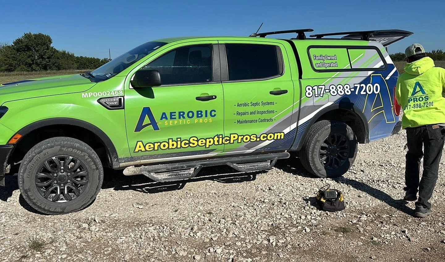 A green and blue company vehicle labeled 'Aerobic Septic Pros' parked on gravel with a person in a neon yellow jacket standing nearby.