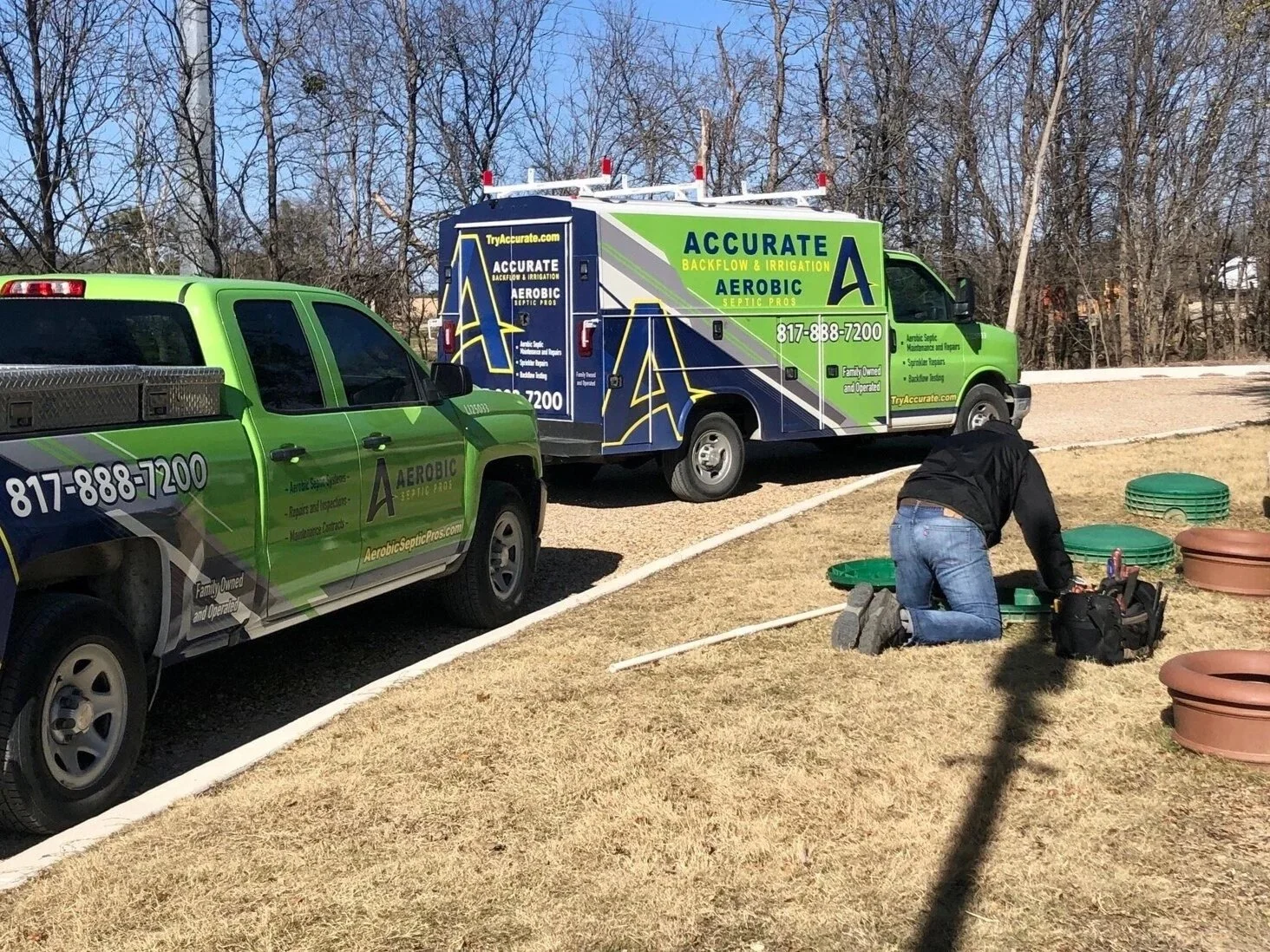 A man kneeling on grass working on plumbing, with two green septic service trucks parked nearby and tree-lined background.