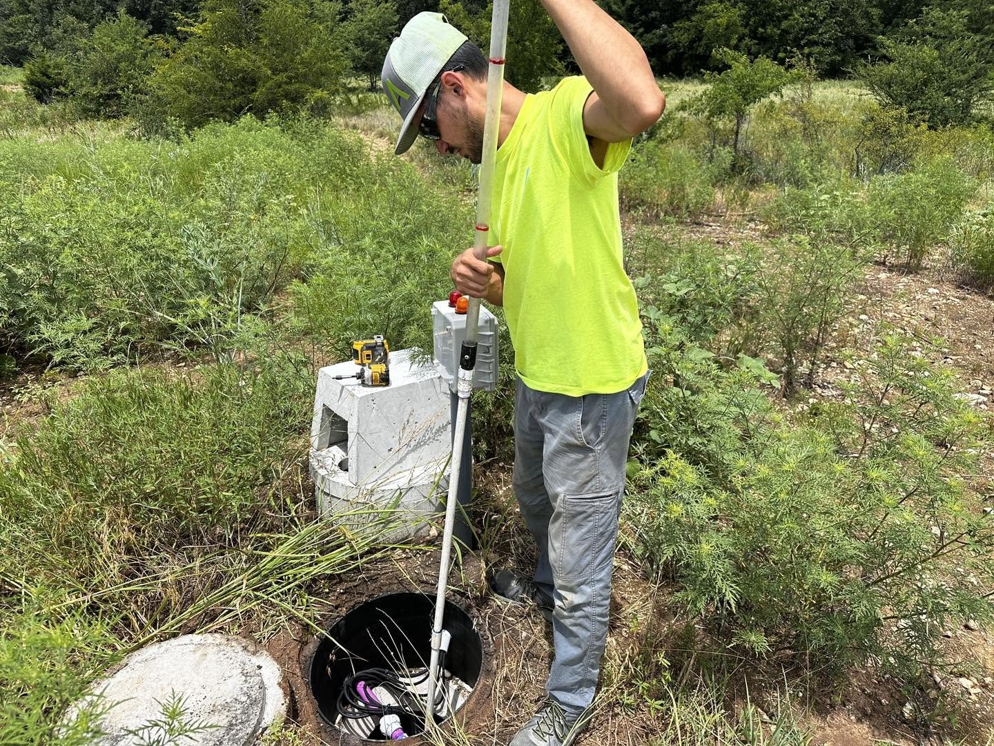 A man in a bright yellow shirt and gray pants inspecting an underground utility access point with equipment, including a drill, a measuring tool, and what appears to be a remote control, outdoors in a green, shrub-filled area.