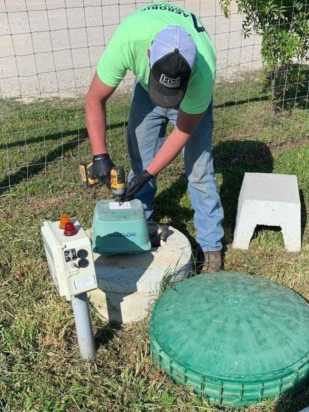 A person wearing a green shirt, jeans, a black cap, and black gloves is working outdoors with a drill on a small machine or device. There are two concrete blocks and a green circular object in the grassy area near a wire fence.