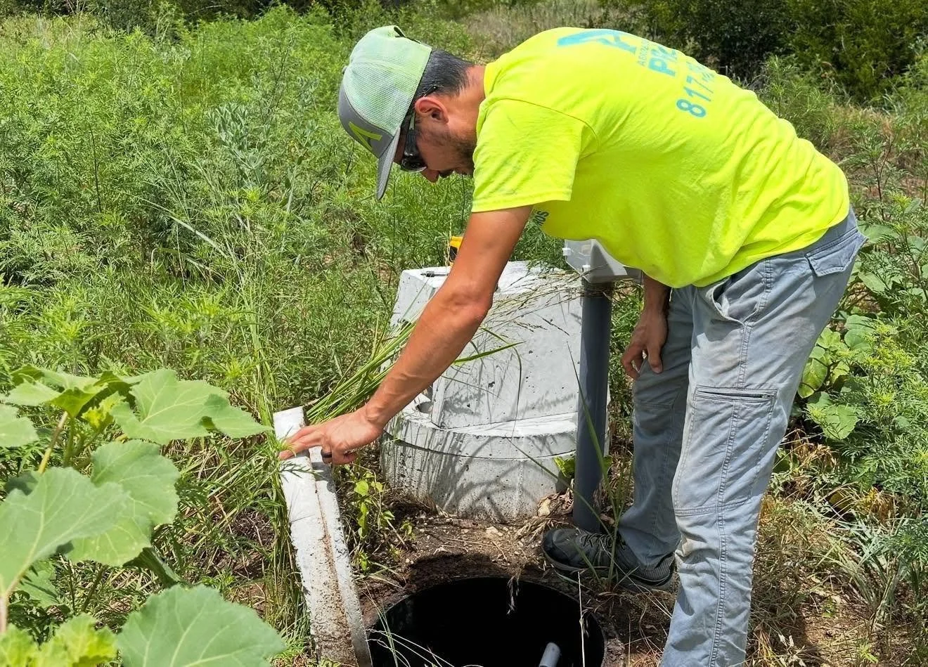 A man in a yellow shirt and gray cap is working outside near a groundwater monitoring well, attaching or adjusting equipment in an overgrown area.