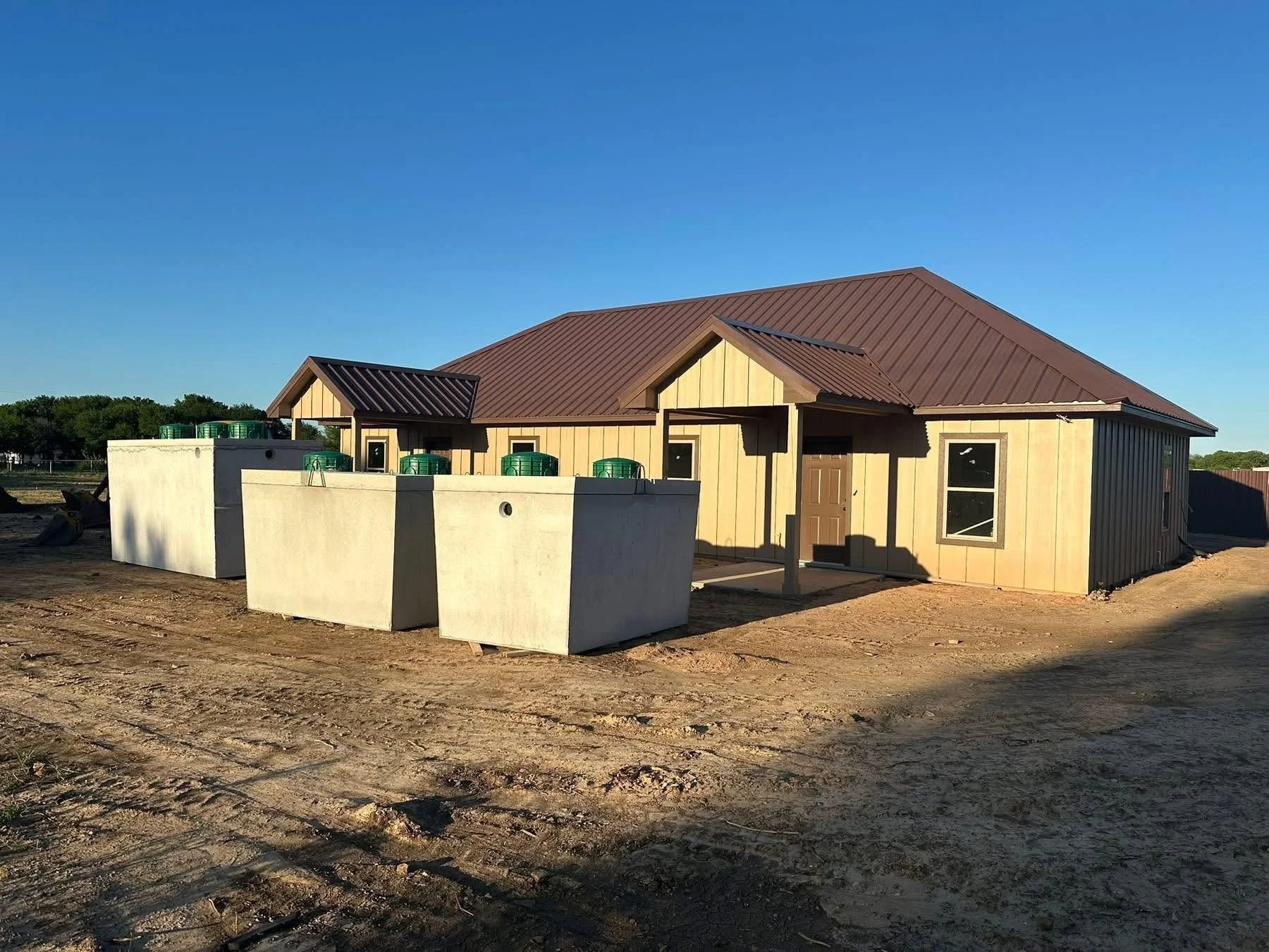 A house under construction with beige and black exterior walls and a brown metal roof, surrounded by construction materials and a dirt lot.