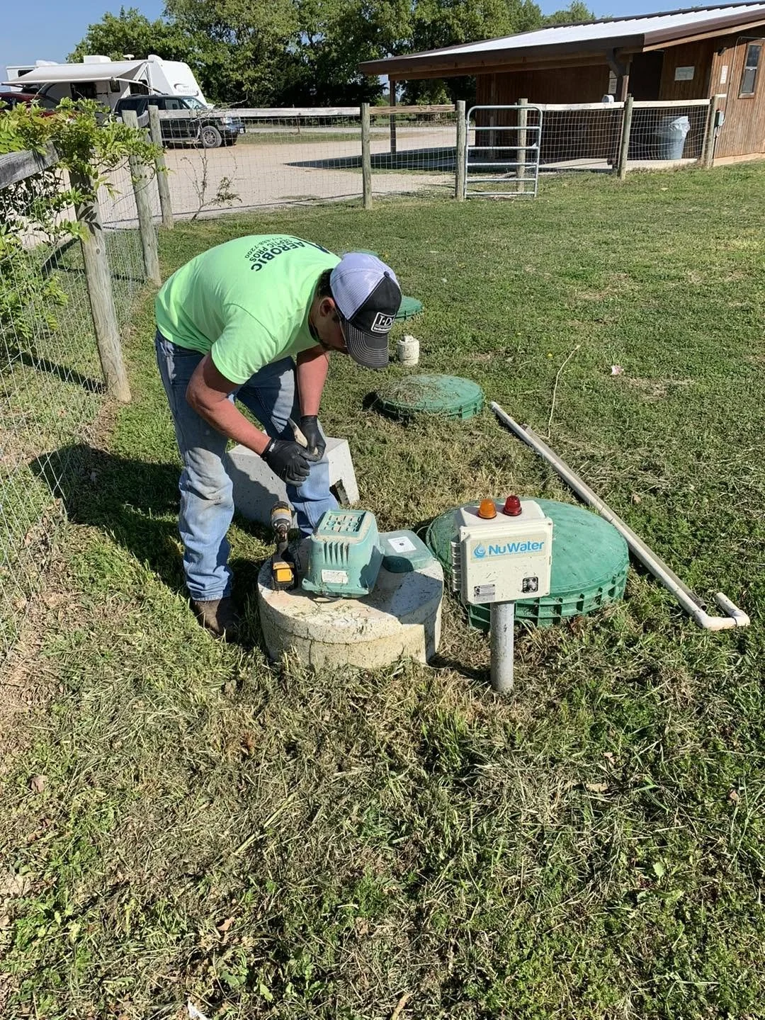 A worker in a green shirt and blue jeans is installing or repairing a water system underground. He is using a drill and is bent over a concrete base near water access points and control boxes labeled "NuWater." There is fencing, a building, and vehic