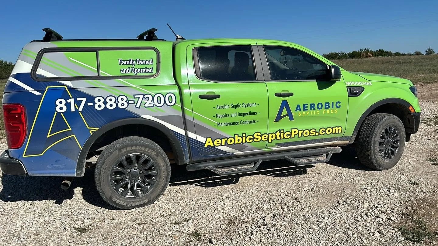 A green and blue pickup truck with decals advertising Aerobic Septic Pros, featuring the company's logo, phone number 817-888-7200, website aerobicsepticipros.com, and services including aerobic septic systems, repairs, inspections, and maintenance.
