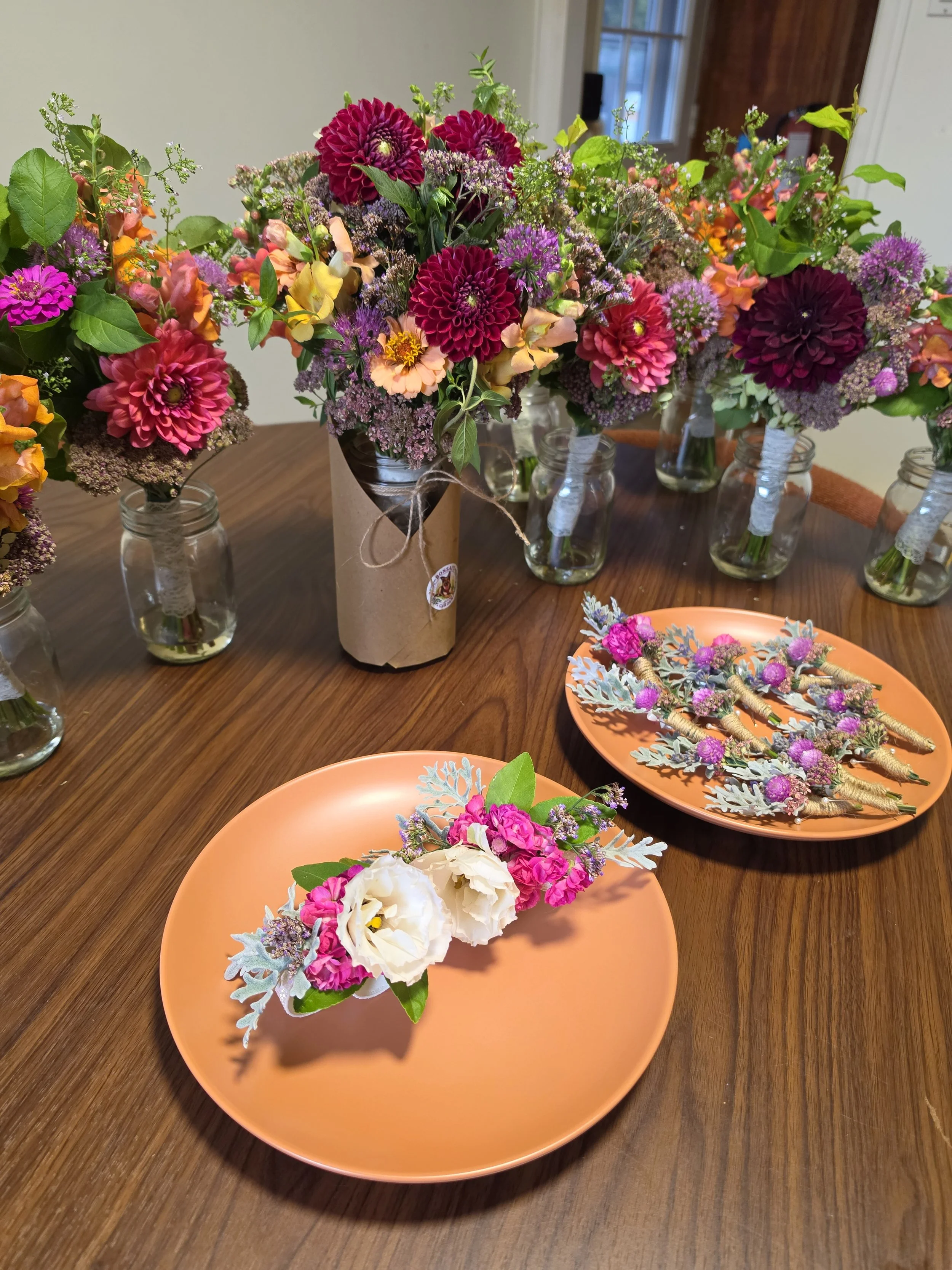 Arrangement of various colorful flowers for weddings (bridal bouquet, bridesmaid bouquets, corsages, boutonnieres) including dahlias, roses, and greenery in glass jars and decorative plates on a wooden table.