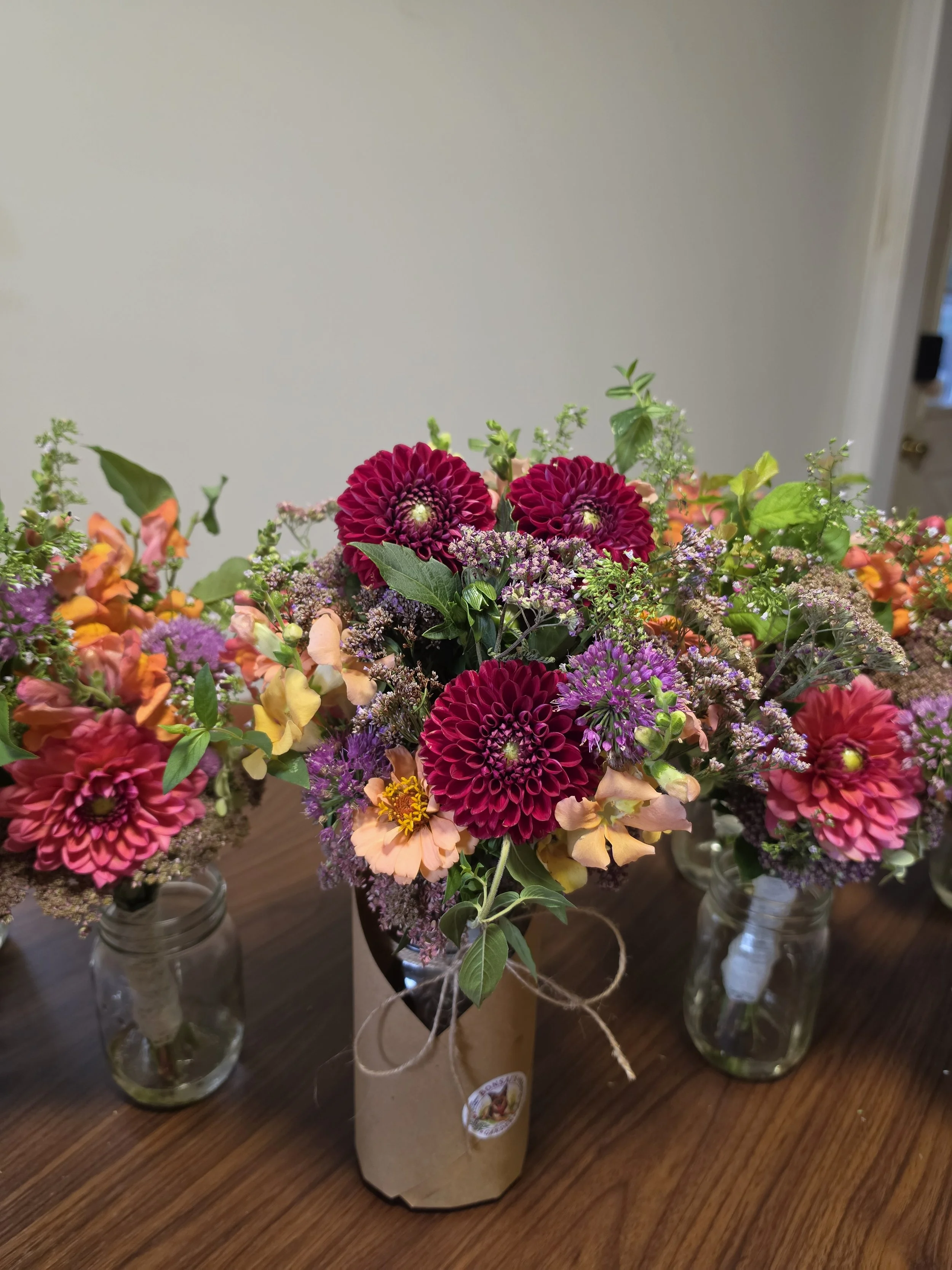 Multiple vases of a bridal bouquet and bridesmaid bouquets, with colorful fresh flowers on a wooden table, including pink dahlias, purple flowers, orange and pink blossoms, and green foliage.