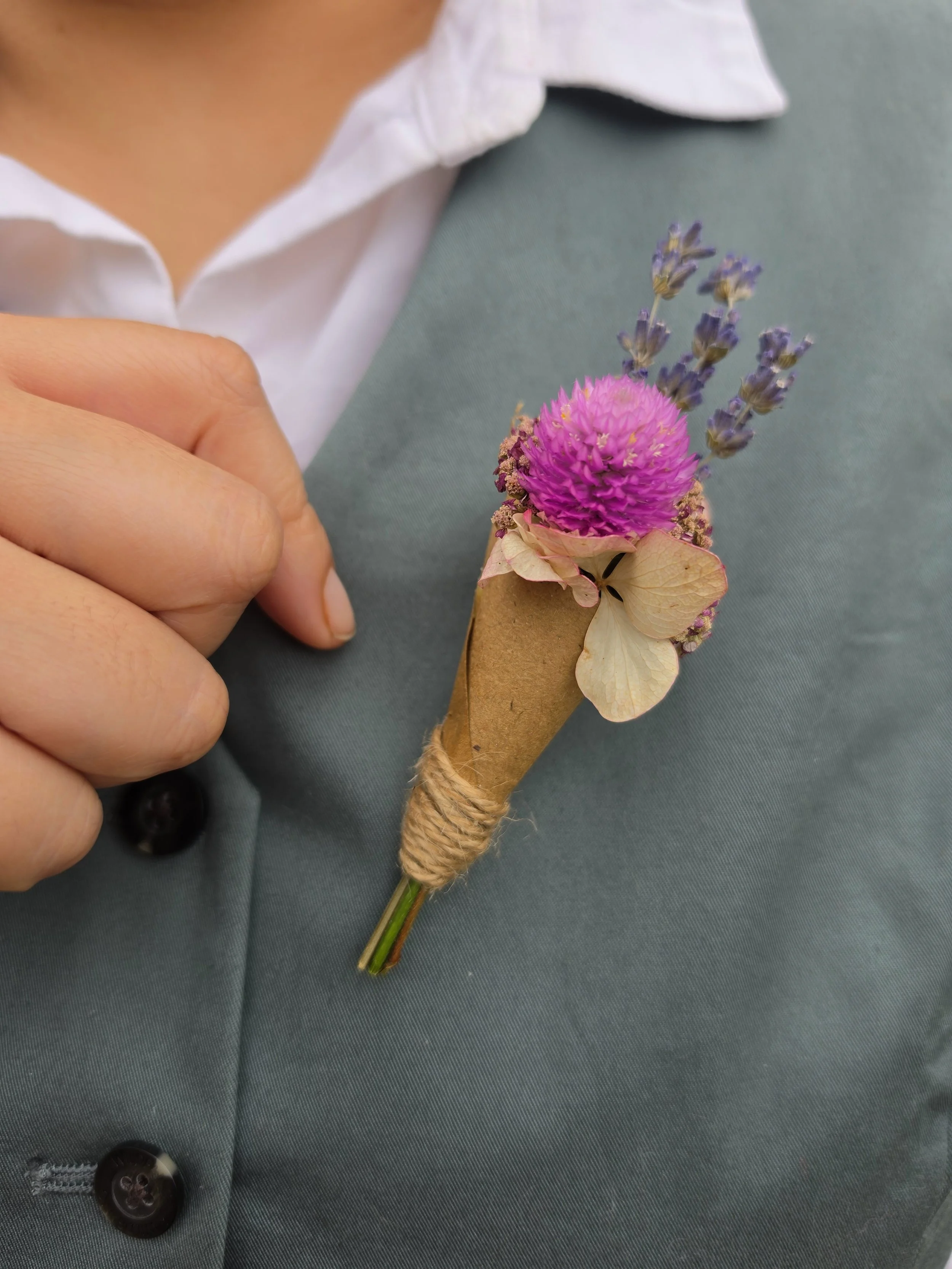 Close-up of a person's hand pinning a small unique crafted groom bouquet boutonniere of flowers with purple and pink blossoms onto a green suit jacket.