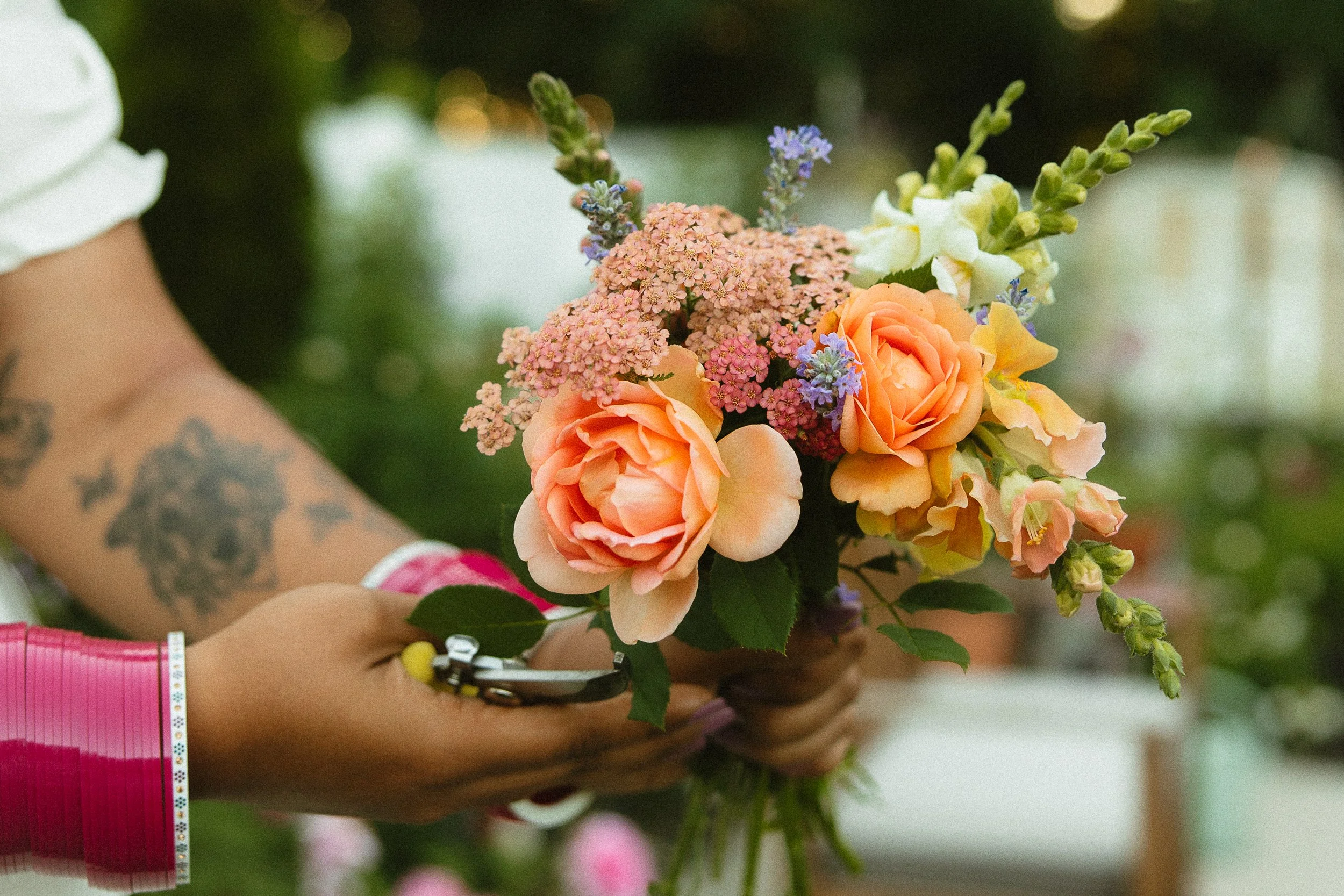 Person holding a locally grown bouquet of assorted flowers, including peach roses, pink yarrow, white snapdragons, and purple lavender, with tattoos visible on arm.