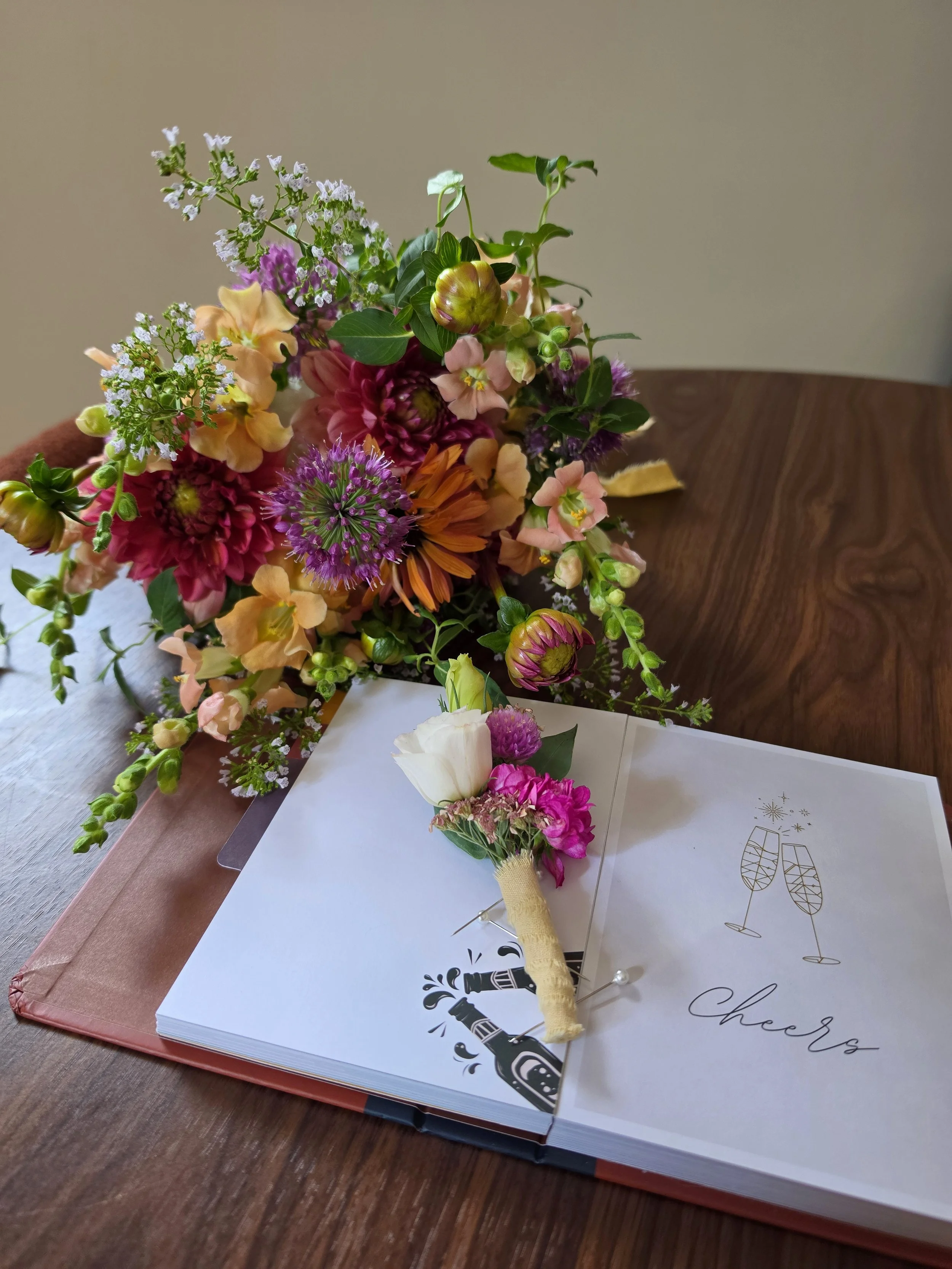 A colorful bouquet of various flowers, including pink, yellow, purple, and white blossoms, arranged on a wooden table along with a wedding or celebration card and a boutonniere.