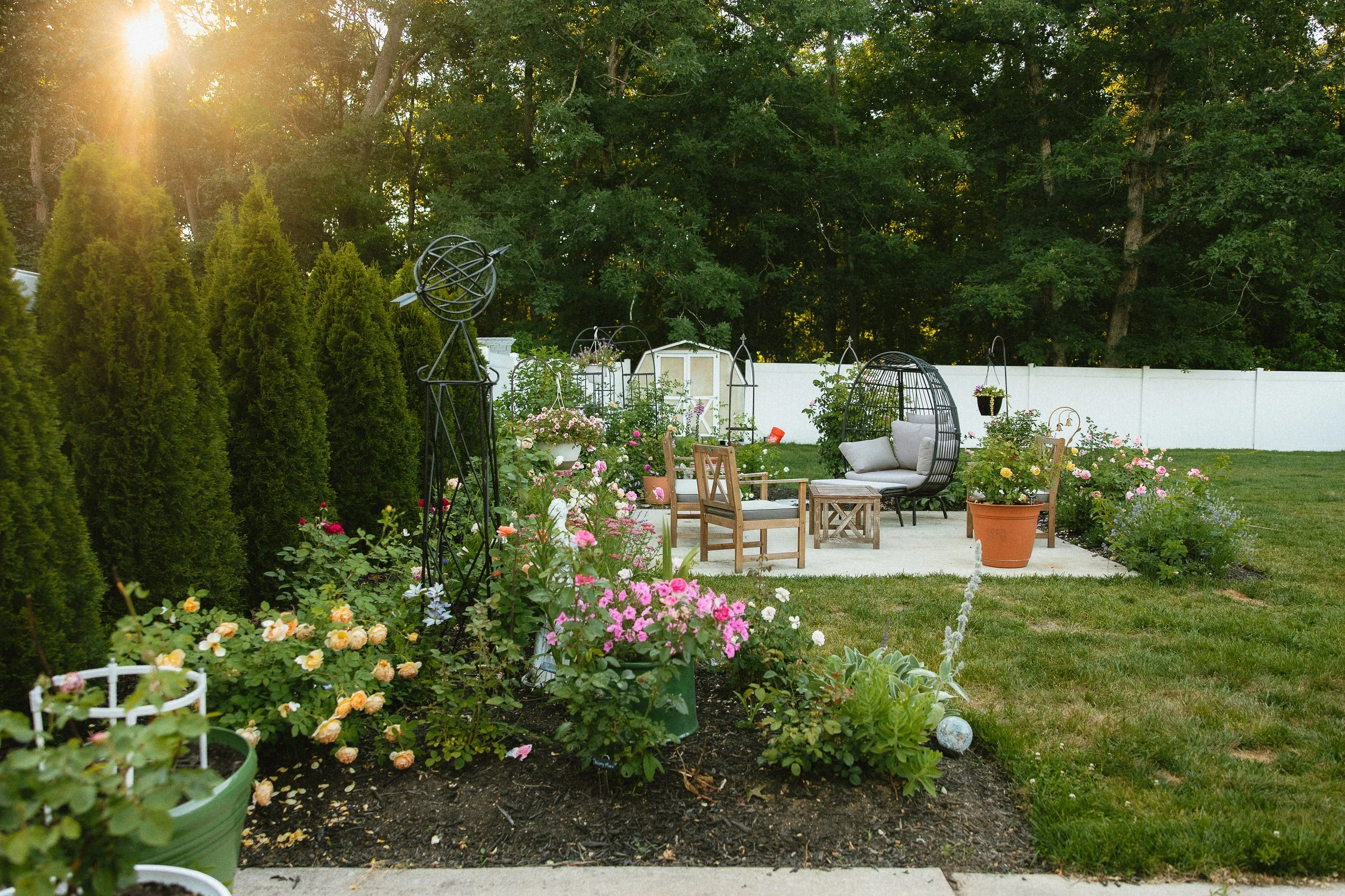 A backyard garden with colorful flowers, potted plants, green shrubs, and outdoor furniture including chairs and a hanging cocoon-style chair, surrounded by a white fence and tall trees in the background with sunshine.
