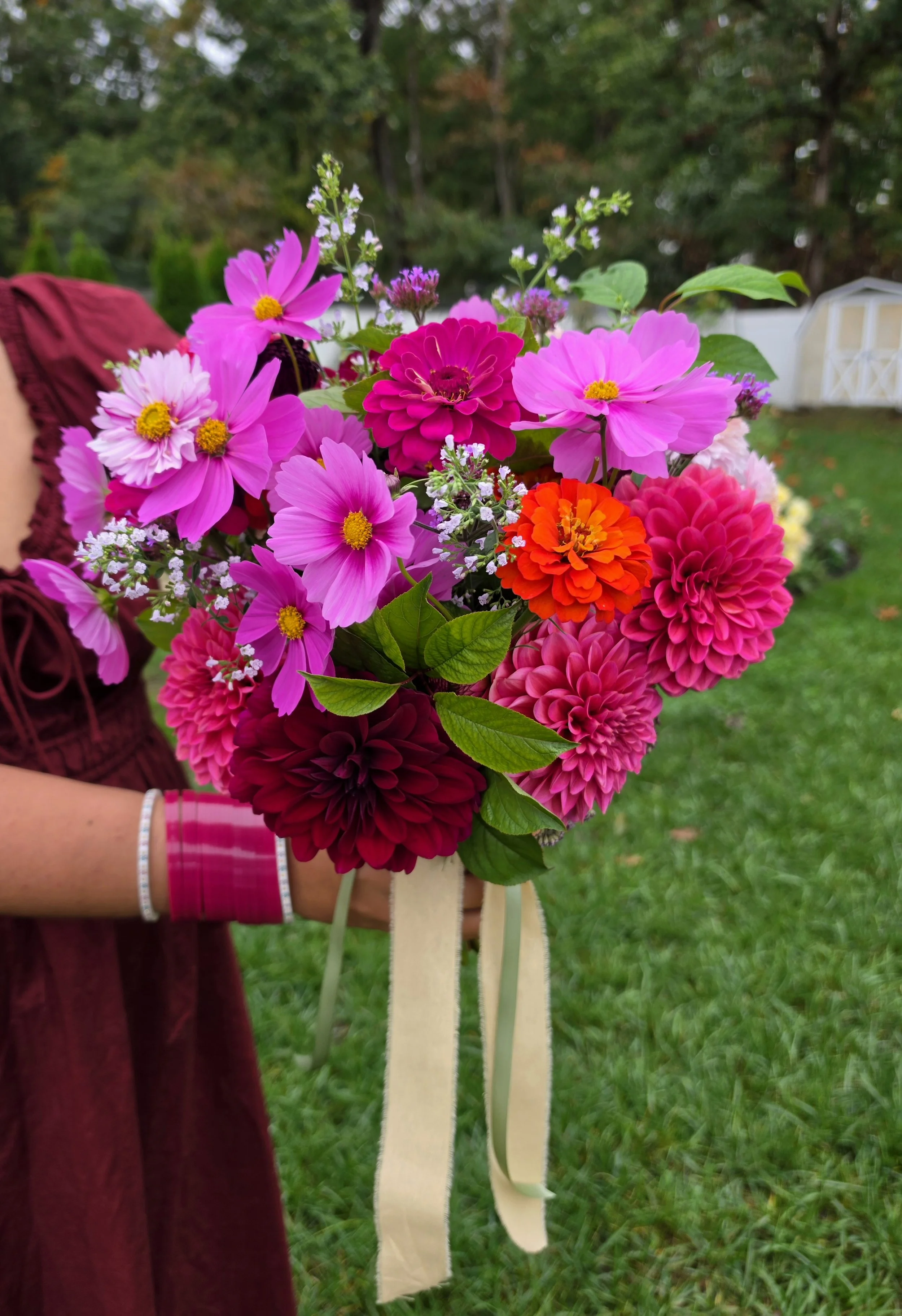 A person holding a large bridal bouquet of vibrant pink, red, and purple flowers outdoors on green grass, with trees and a white structure in the background.