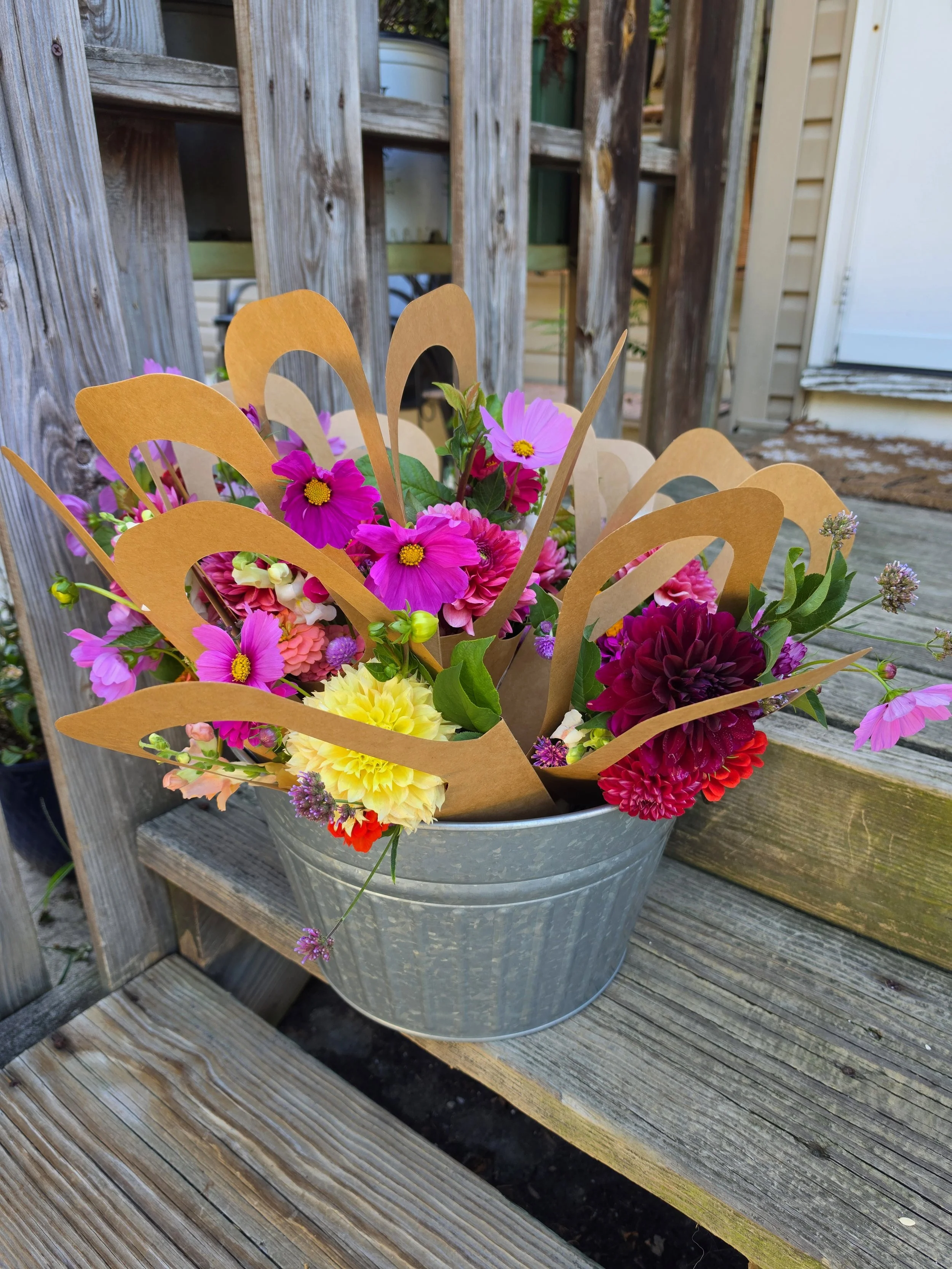 A metal bucket filled with colorful pink, yellow, and purple flowers, with some decorative brown paper around them for bloom bucket events.
