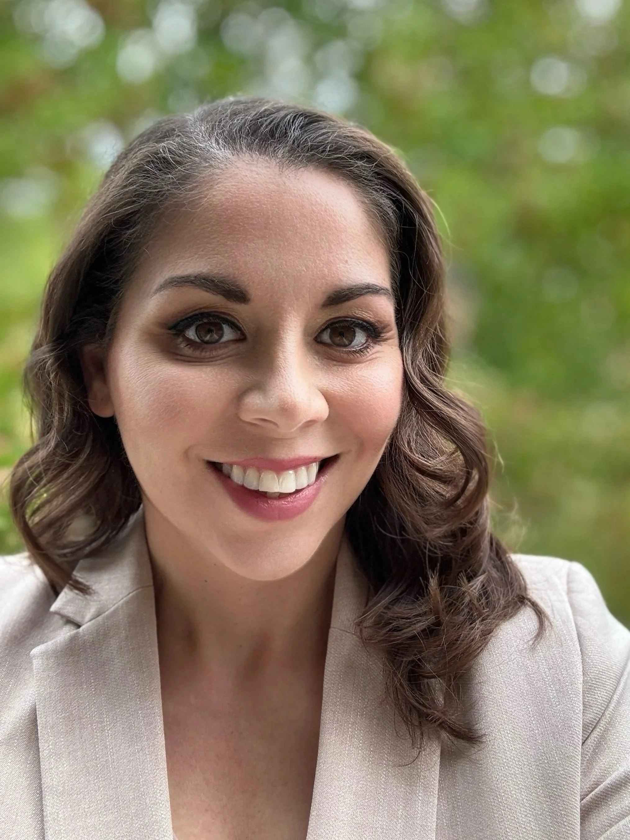 Close-up of a smiling woman with long, curly brown hair, wearing a beige blazer, outdoors with a blurred green background.