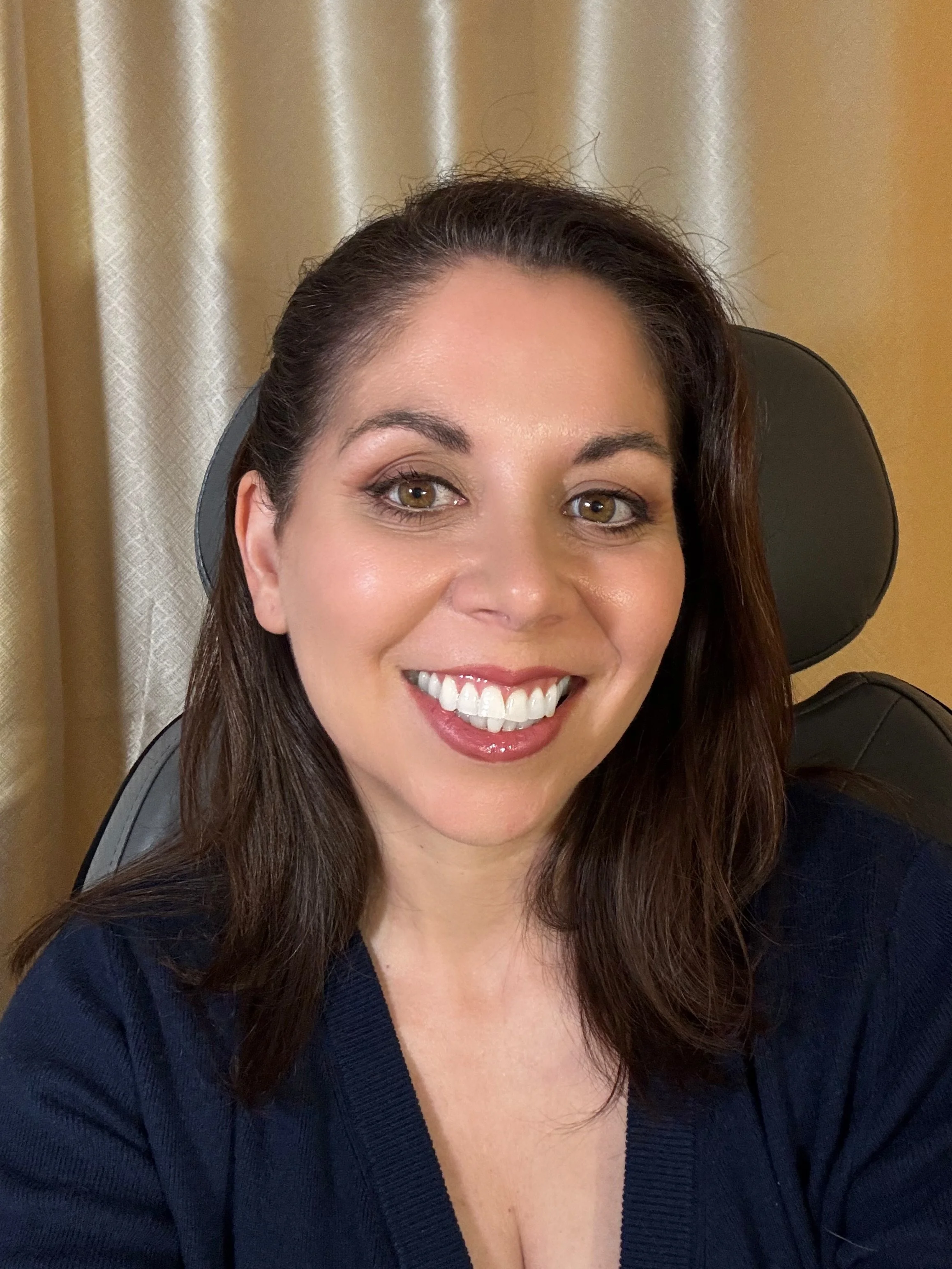 A smiling woman with dark brown hair sitting on a black chair in front of a beige curtain, wearing a dark blue top.