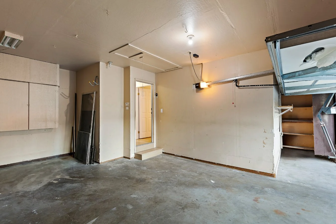 Empty garage with concrete floor, white walls, and an open garage door with a fish tank on top. There is a doorway to a room with hardwood flooring and a white door inside. Storage cabinet and tools lean against the wall.