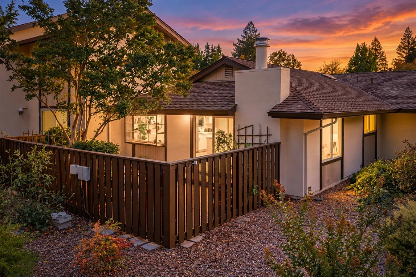 A cozy house with a deck surrounded by garden, lit from inside during sunset, with a colorful sky, trees, and a chimney.