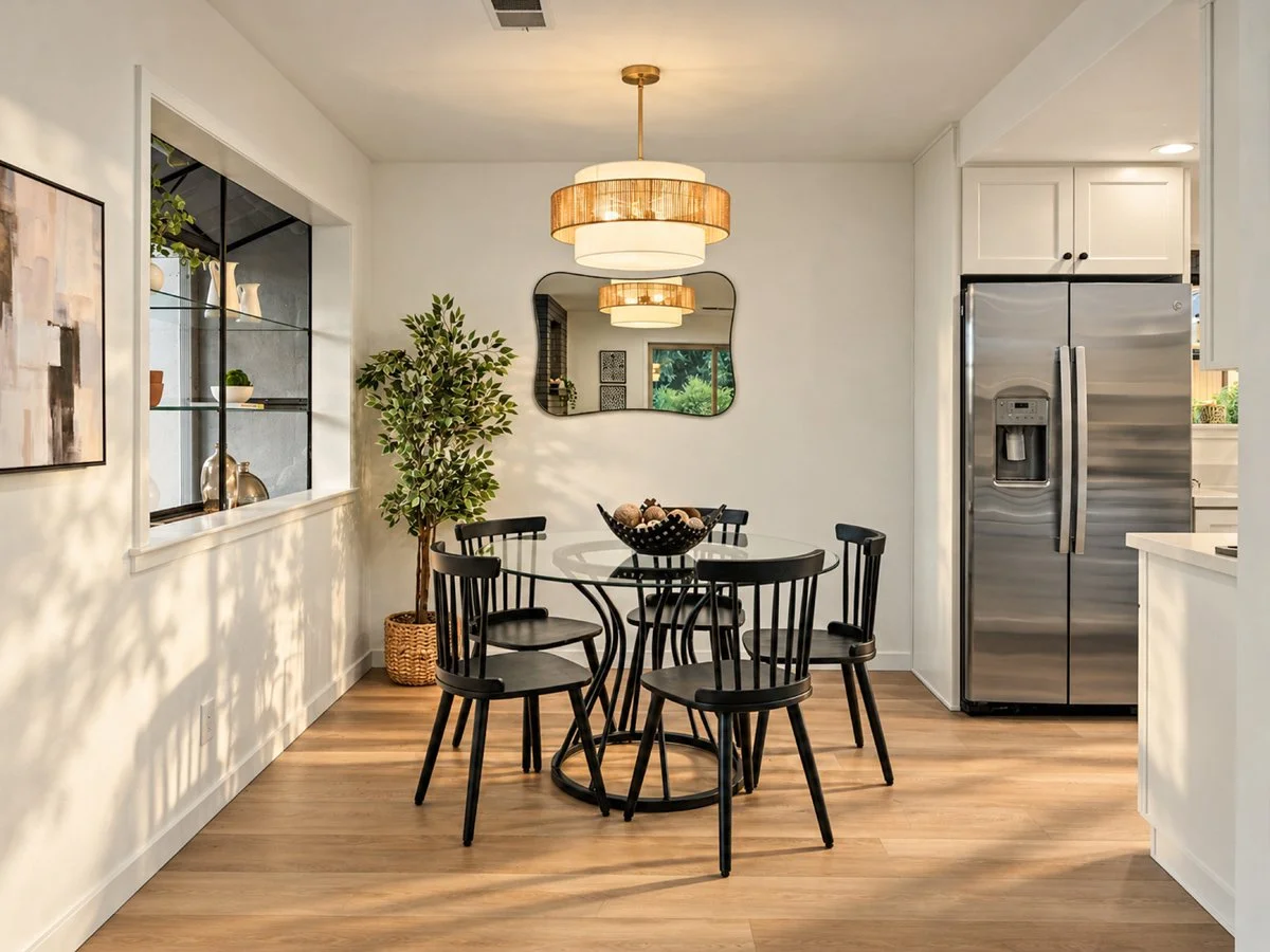 Dining area with a glass table surrounded by six black chairs, large mirror on white wall, modern chandelier overhead, potted plant in corner, open kitchen with stainless steel refrigerator to the right, and decorative items on window shelf.