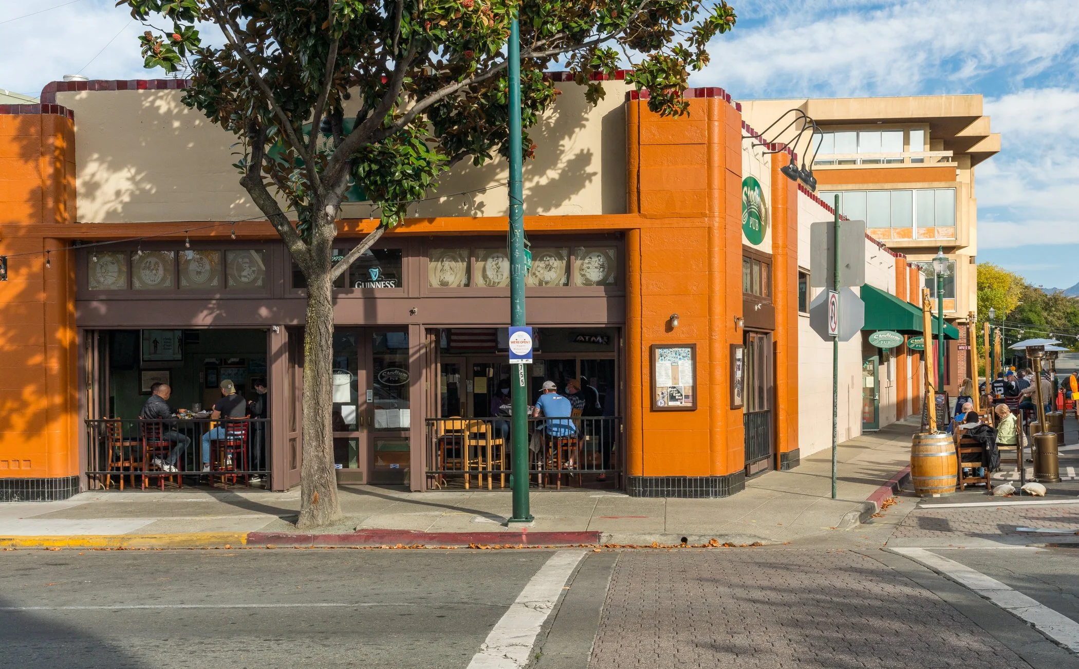 A lively street scene featuring a building housing a pub with a green sign, outdoor seating with people dining, and a tree in front. The weather is sunny with some clouds in the sky.