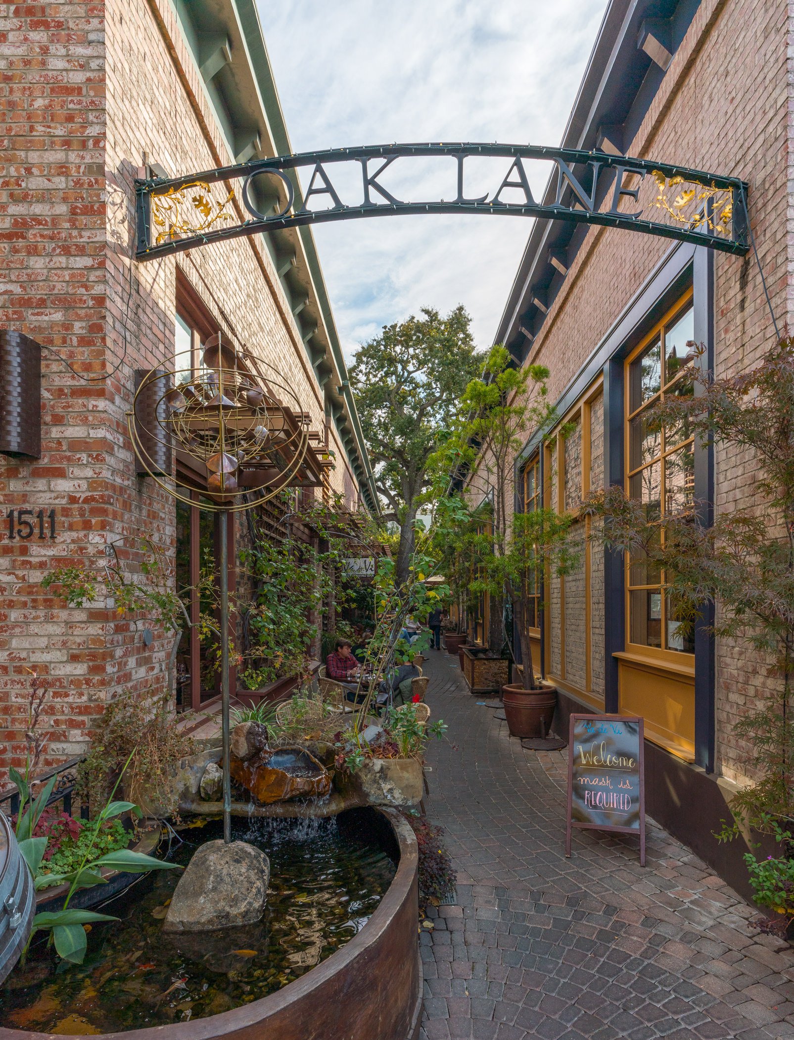 An alleyway scene with brick buildings on either side, a water fountain with rocks and plants, and a small sign that reads 'Welcome, mask is required'.