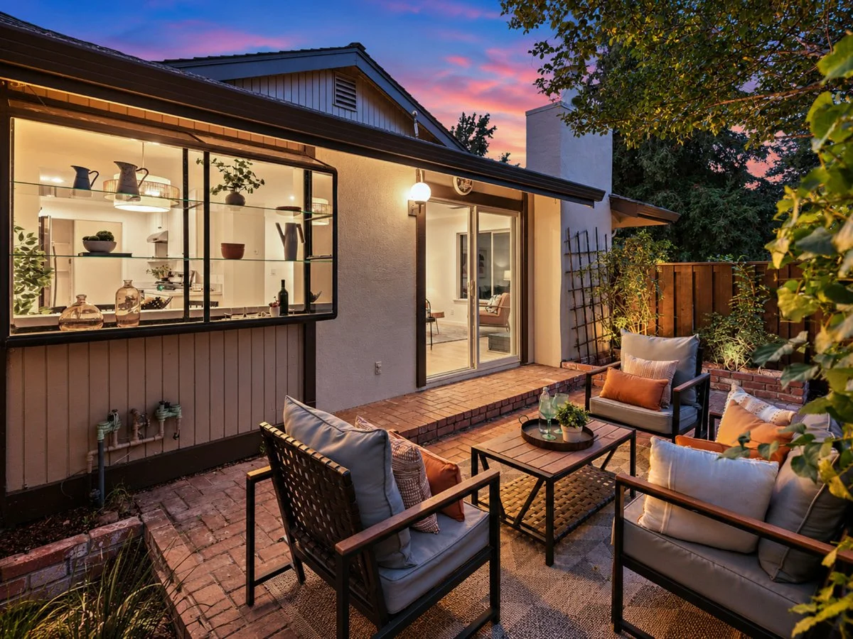 Cozy outdoor patio with seating area, pillows, coffee table, and lush greenery, illuminated at dusk with the house interior visible through sliding glass doors.