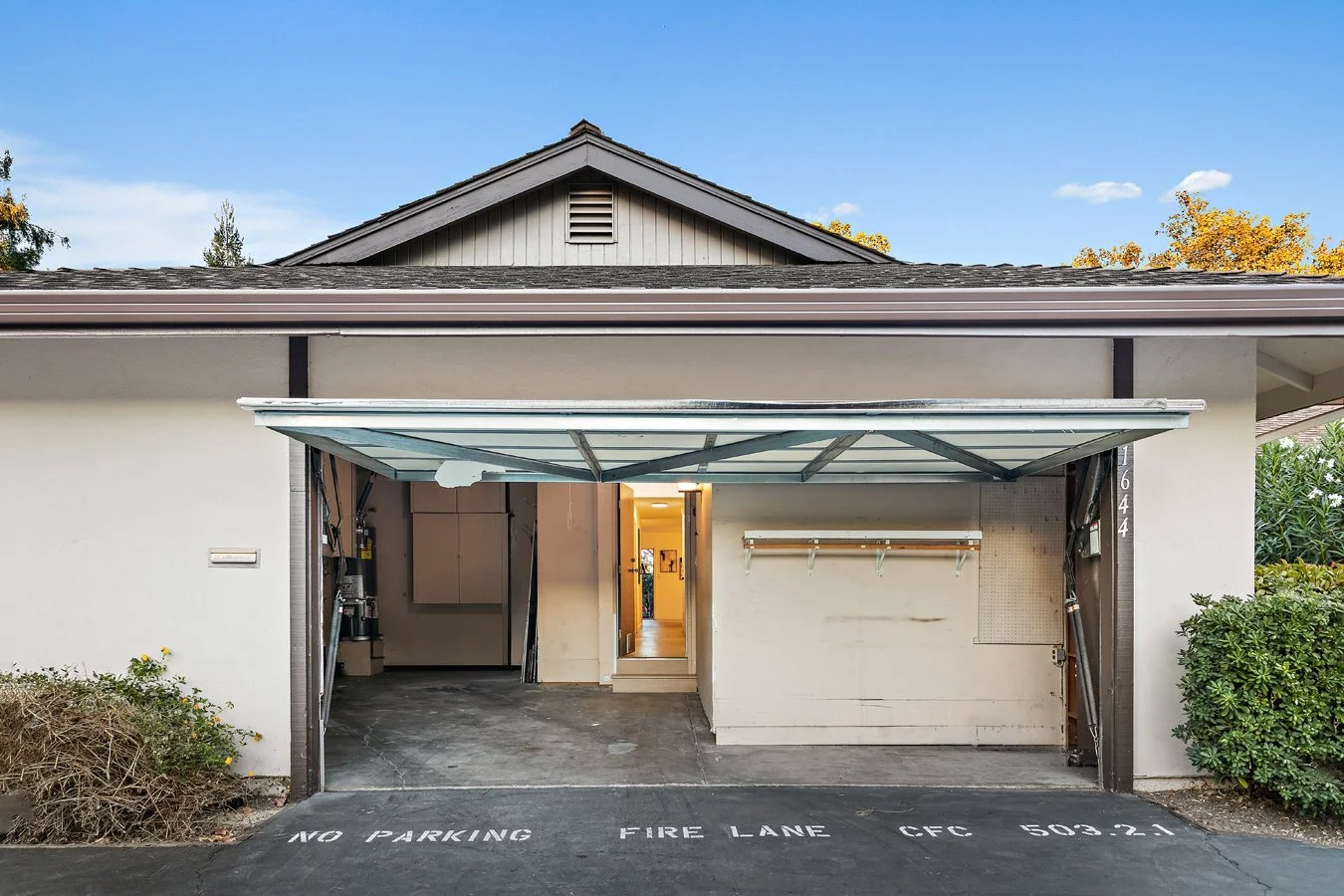 View of a residential garage with an open door, showing a bike rack on the wall inside. The garage is attached to a house, with a visible stairway leading inside. The driveway has painted parking restrictions, and the sky is partly cloudy with some t