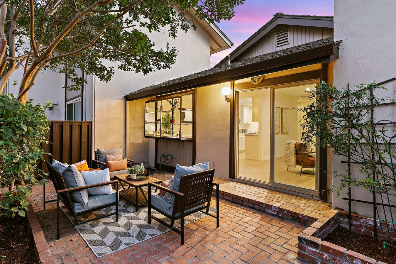 A cozy backyard patio with outdoor seating, a small brick step, and views into a well-lit living room through sliding glass doors.