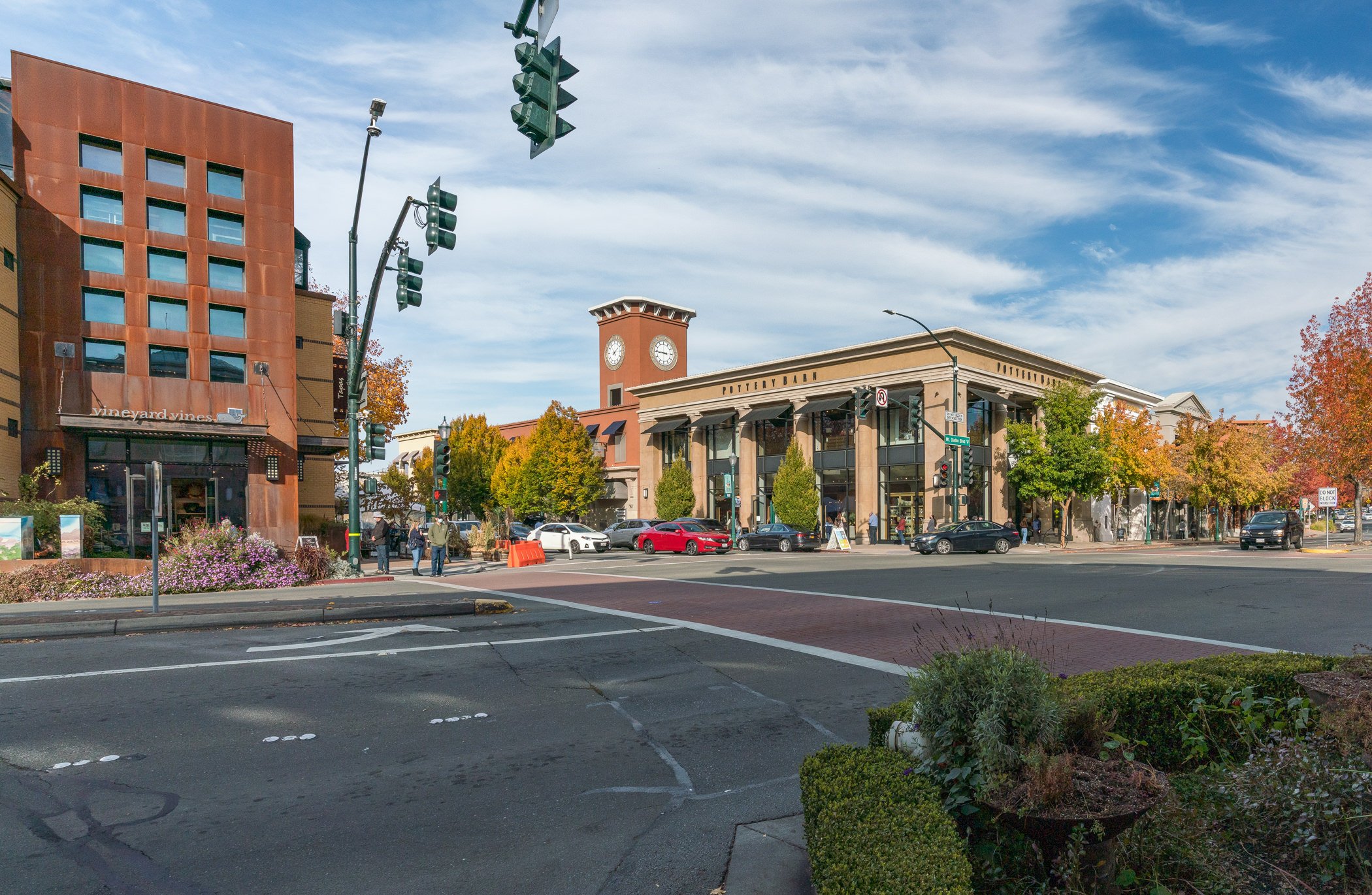 Street scene with buildings, traffic lights, cars, trees with fall foliage, and pedestrians on a sunny day.
