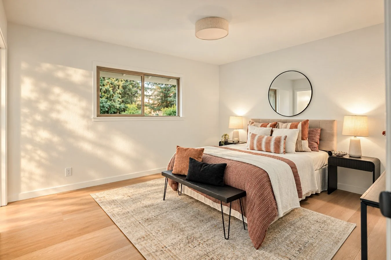 Bedroom with a bed dressed in white and rust-colored bedding, black bench at foot, nightstands with lamps, window showing greenery, round mirror above bed, wooden floors, neutral wall color.