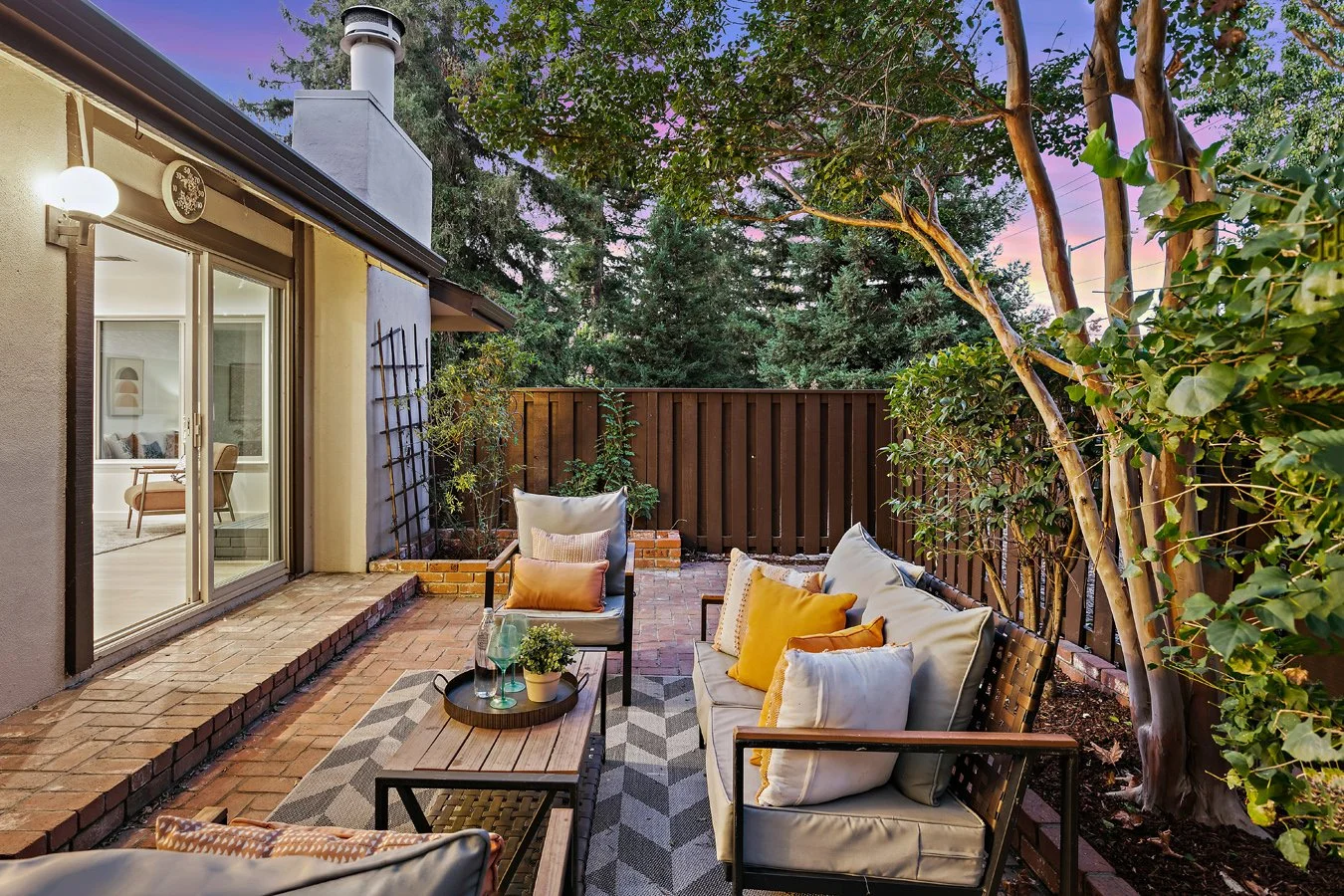 Cozy backyard patio with cushioned outdoor seating, a coffee table with drinks and plants, surrounded by greenery and enclosed with a wooden fence at sunset.