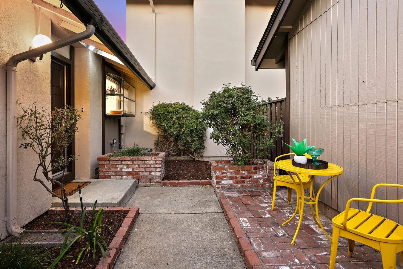 Small outdoor patio with brick borders, yellow metal chairs, green potted plants, shrubs, and a brick planter, enclosed by beige and gray walls.