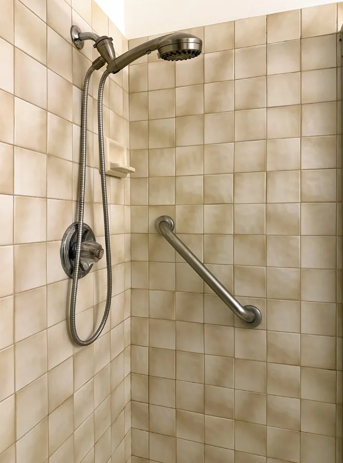 A shower with beige square tiles, a handheld showerhead mounted on a sliding bar, a metal grab bar on the wall, and a small soap dish.