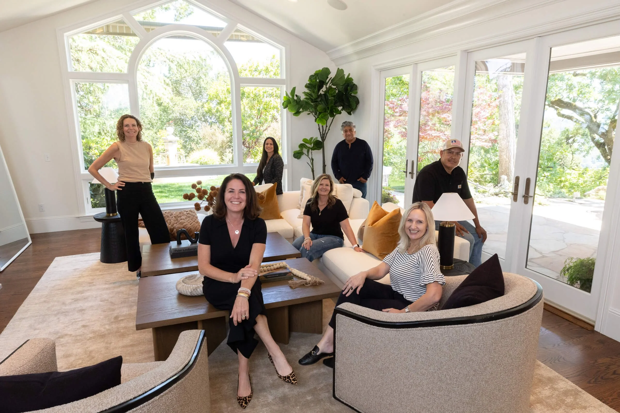 A group of seven people in a bright, modern living room with large windows and outdoor views. Four women are sitting on a sofa and chairs, and three men are standing behind them, smiling and posing for a photo.