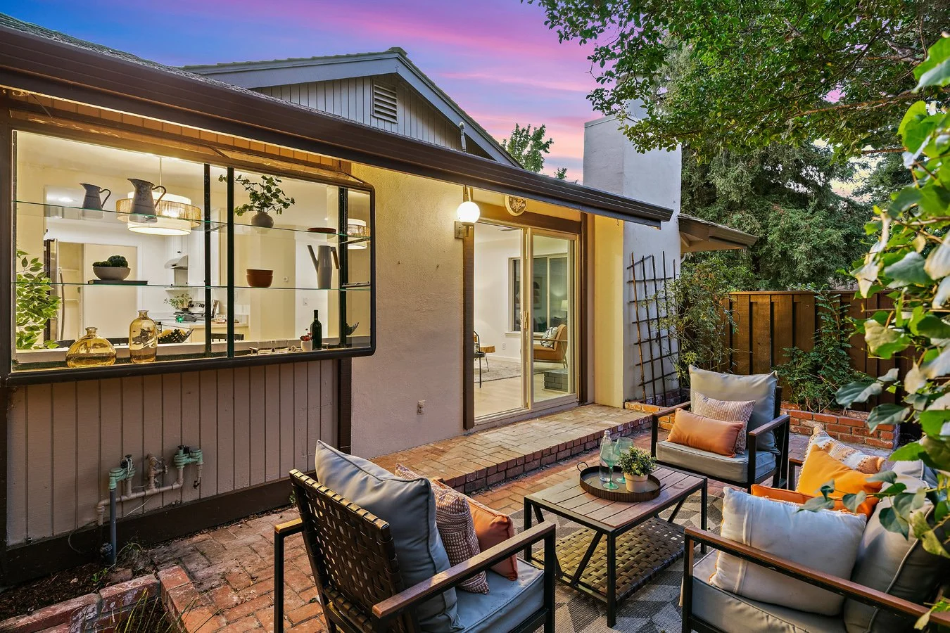 Outdoor patio at sunset with seating area and view into the house kitchen through large window.