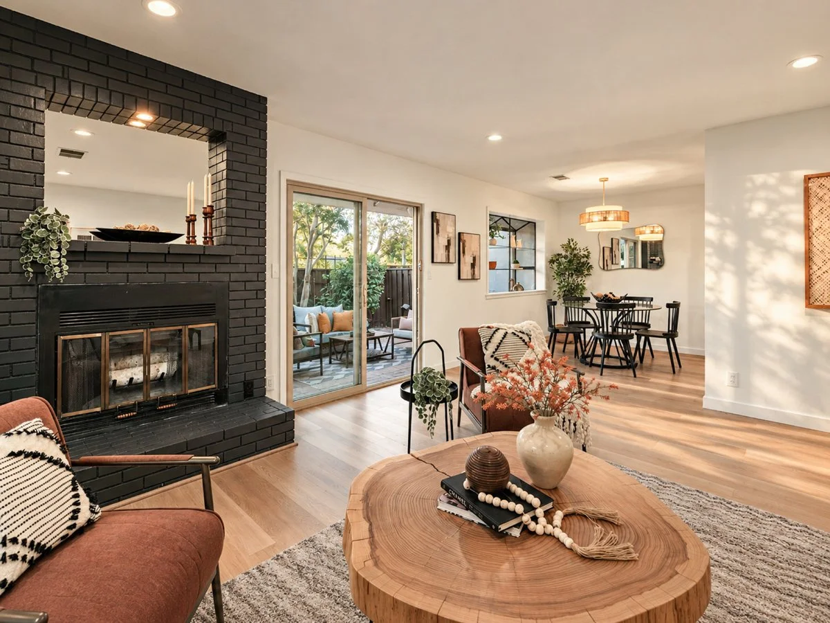 Living room with black brick fireplace, sliding glass door leading to patio, round wooden coffee table with vase and decorative items, pink and black armchair, dining area with round table and black chairs, artwork on walls, and large window with gre