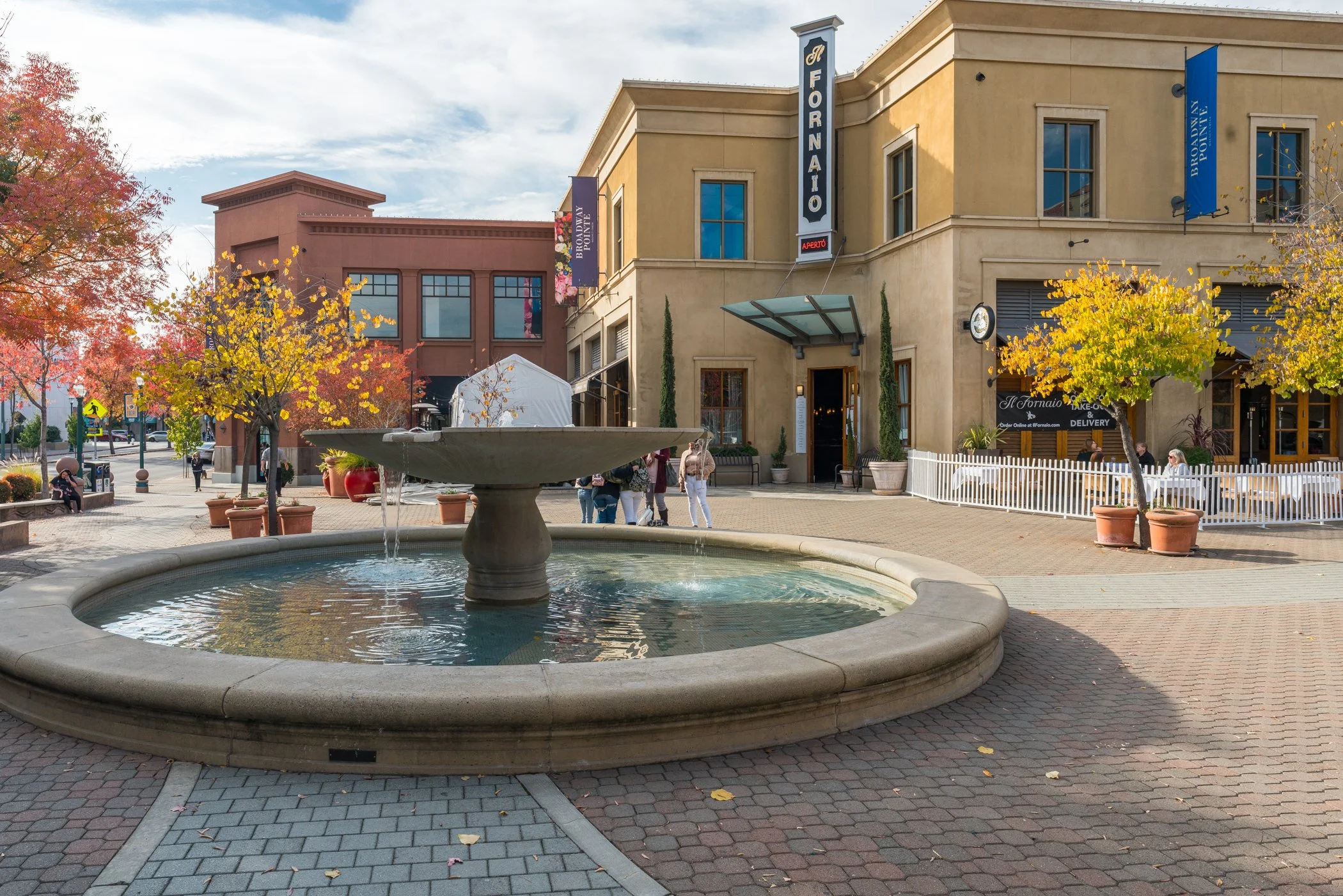 A small town square with a fountain in the foreground, surrounded by trees with autumn foliage. There are buildings with signs that read 'Fornai,' 'Brooklyn Point,' and 'BBQ.' People are walking and sitting around the area.