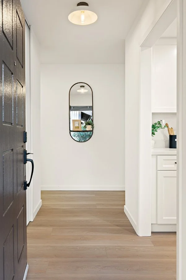 View of a minimalist entryway with light wood flooring, a white wall, a black-framed wall mirror, and a dark front door with black hardware. Part of a white kitchen with a countertop and green plant is visible on the right.