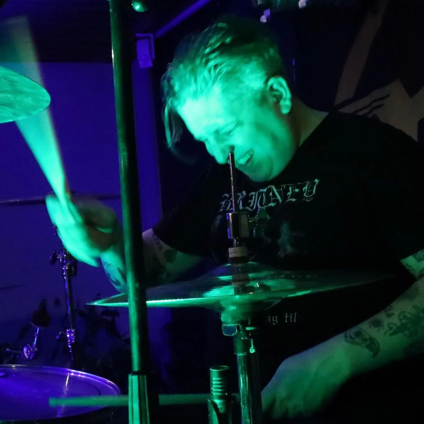 A person with short hair and tattoos on their arm playing drums in a dimly lit setting with green and blue lighting.