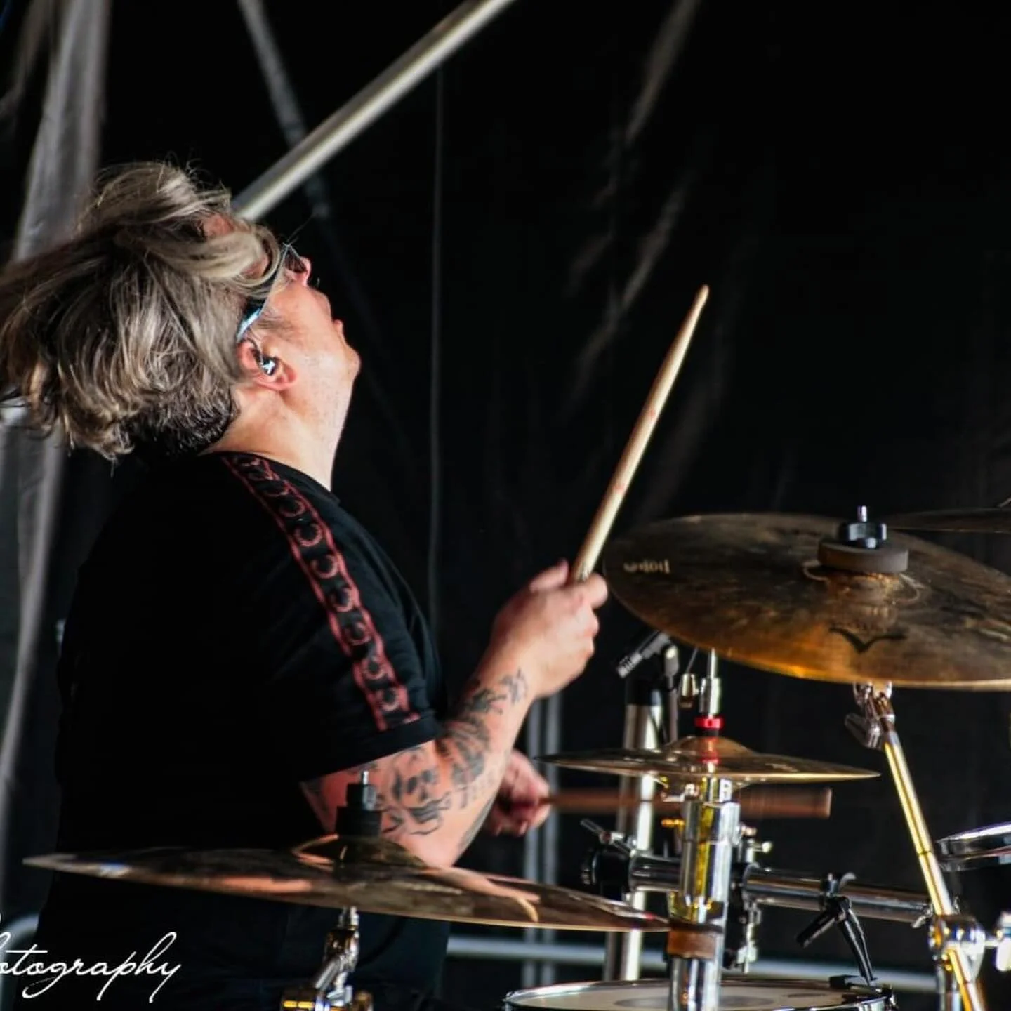 A musician with tattoos on his arms playing drums on stage, wearing glasses and a black shirt with red trim, and a dark background.