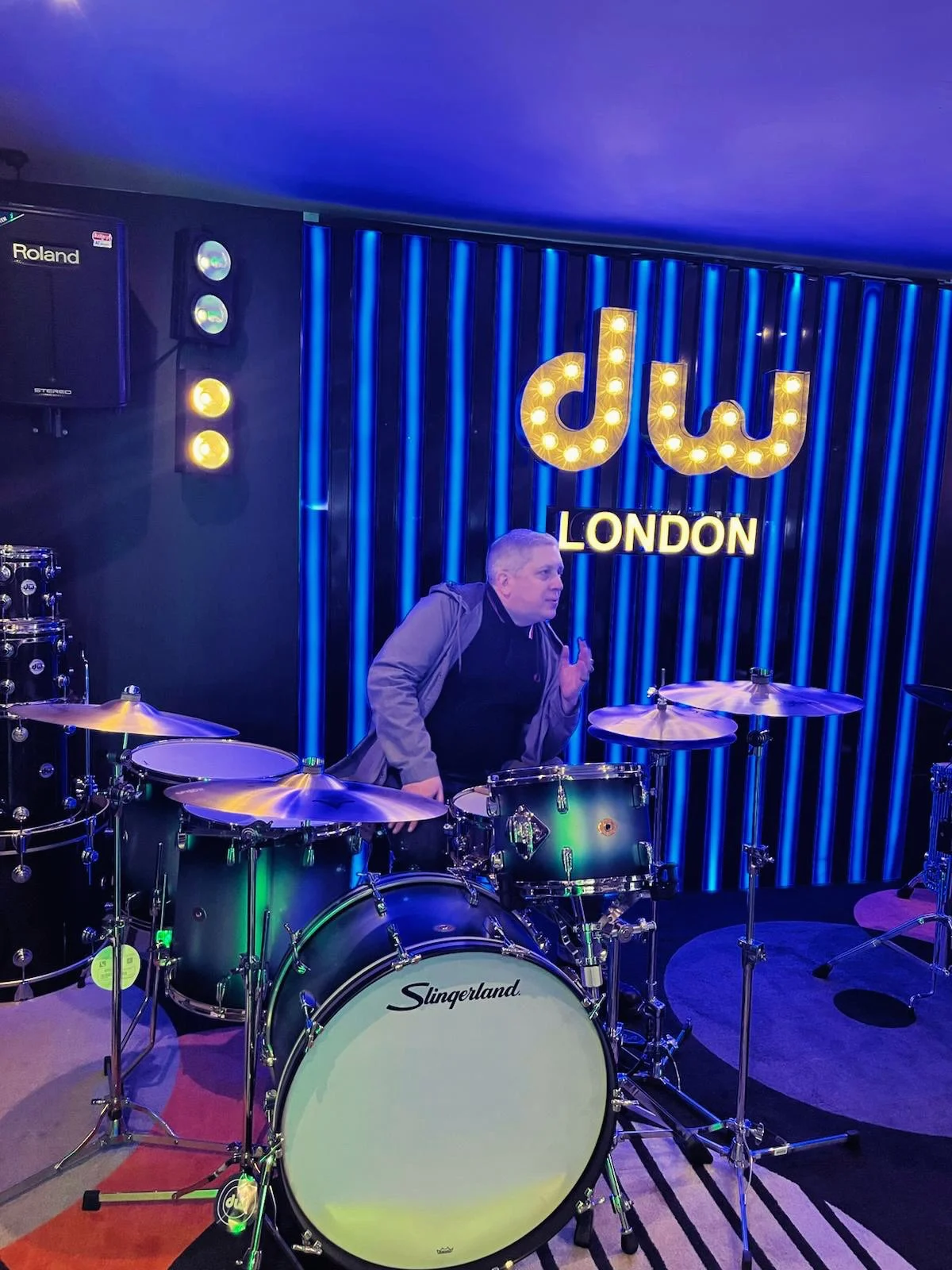 A man standing behind a green Slingeland drum set in a music venue with blue vertical lights and a bright sign reading 'dwt' and 'LONDON' in the background.