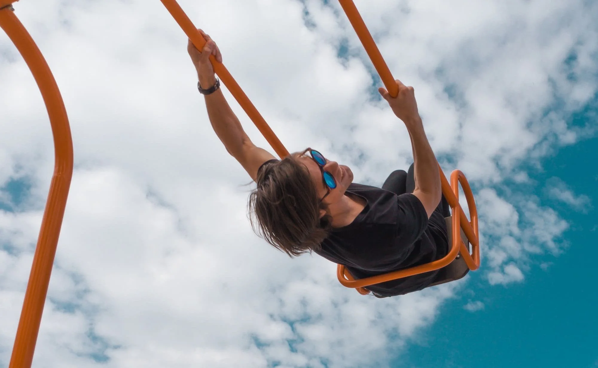 Woman wearing sunglasses riding an orange amusement park swing against a cloudy blue sky