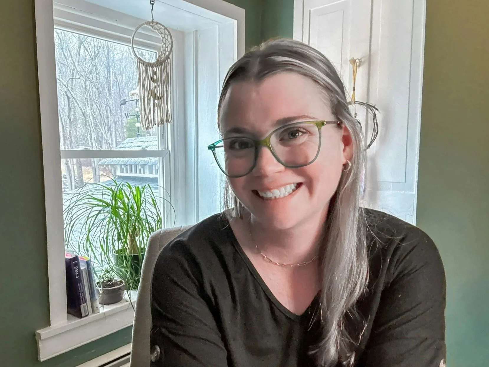 Smiling woman wearing glasses and a black shirt seated indoors beside a window with a potted plant