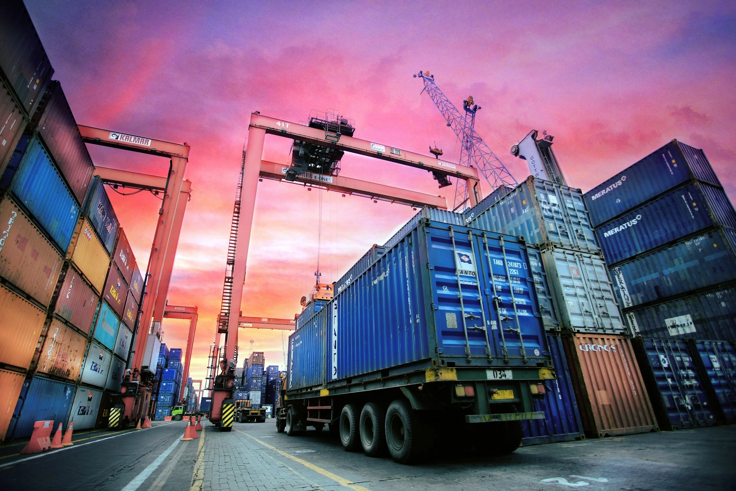 Truck carrying cargo containers under a gantry crane at sunset