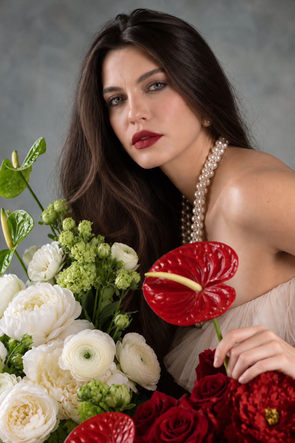 Portrait of a woman with long dark hair, pearl necklace, and red lipstick holding a lush bouquet of white and red flowers.
