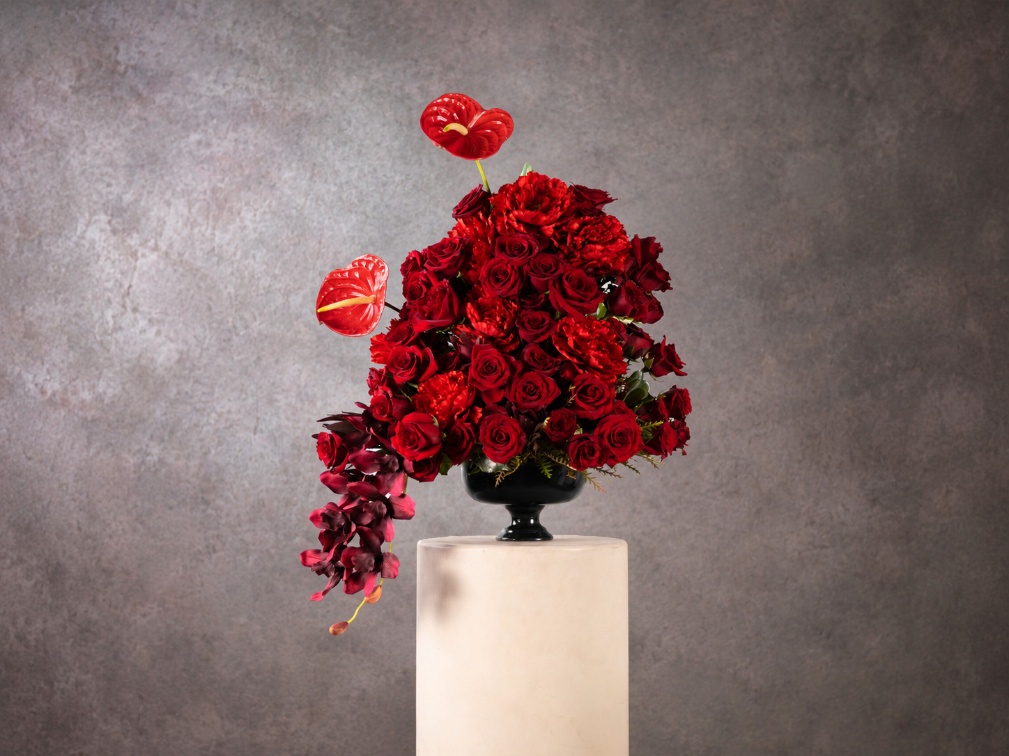 Luxurious arrangement of red roses, carnations, anthuriums, and orchids in a black urn on a white pedestal