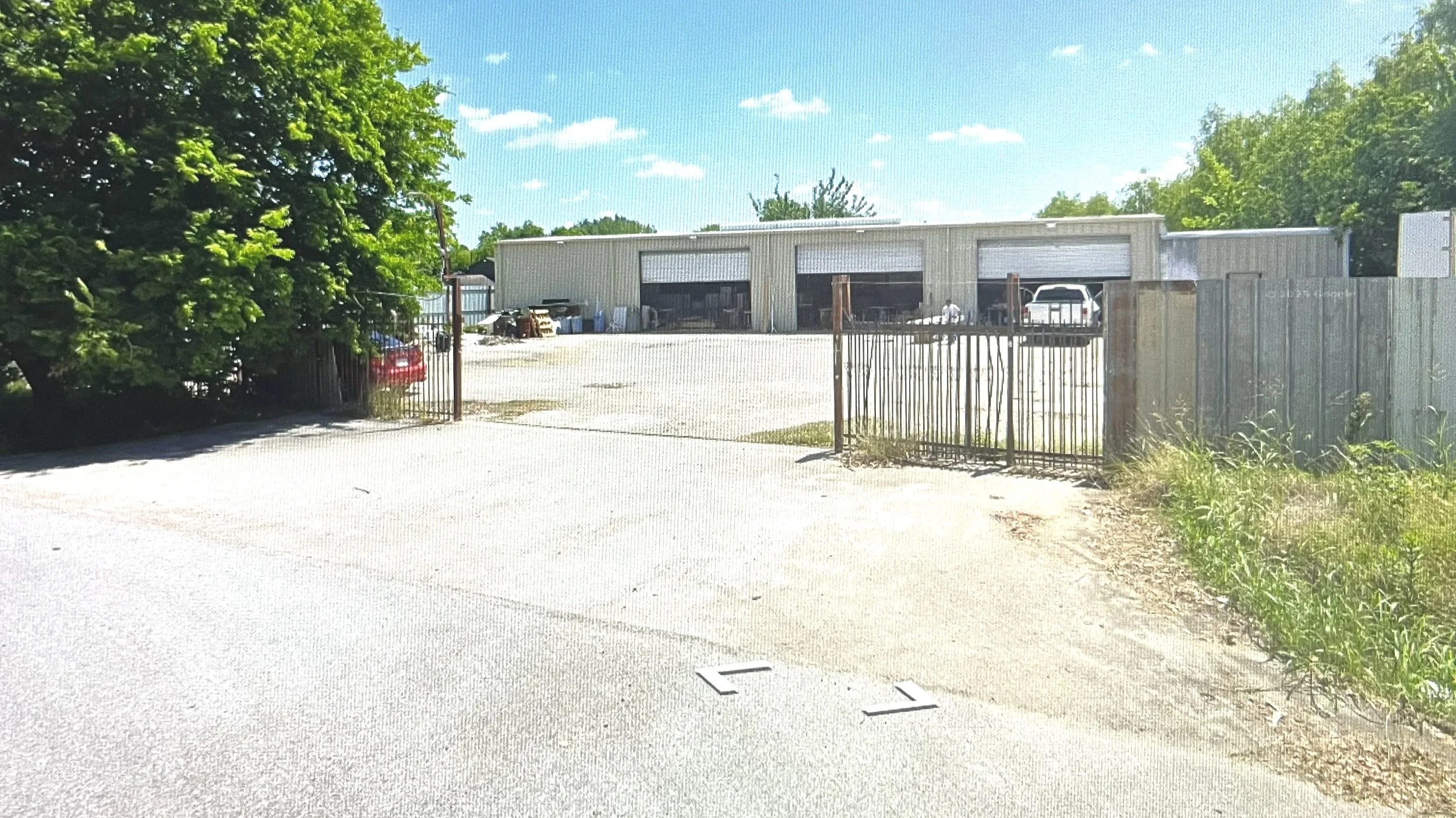 A metal industrial building with three open garage doors, surrounded by a metal fence with a gate. There are trees on either side and a clear blue sky with some clouds.